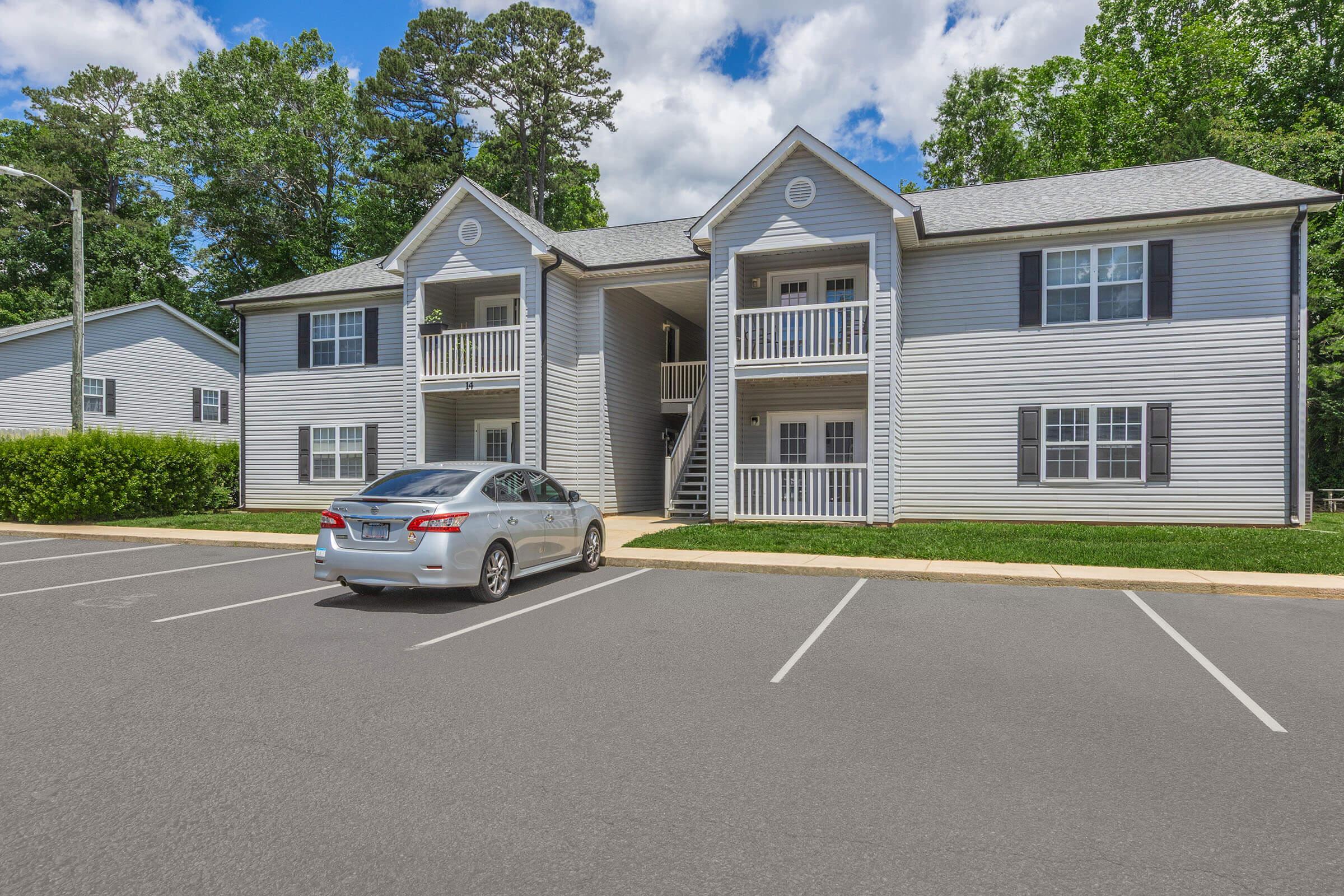 A two-story residential building with a light gray exterior featuring multiple balconies. In the foreground, a silver car is parked in an empty lot. Lush green trees and shrubs surround the property, and the sky is partly cloudy. The area appears well-maintained and inviting.