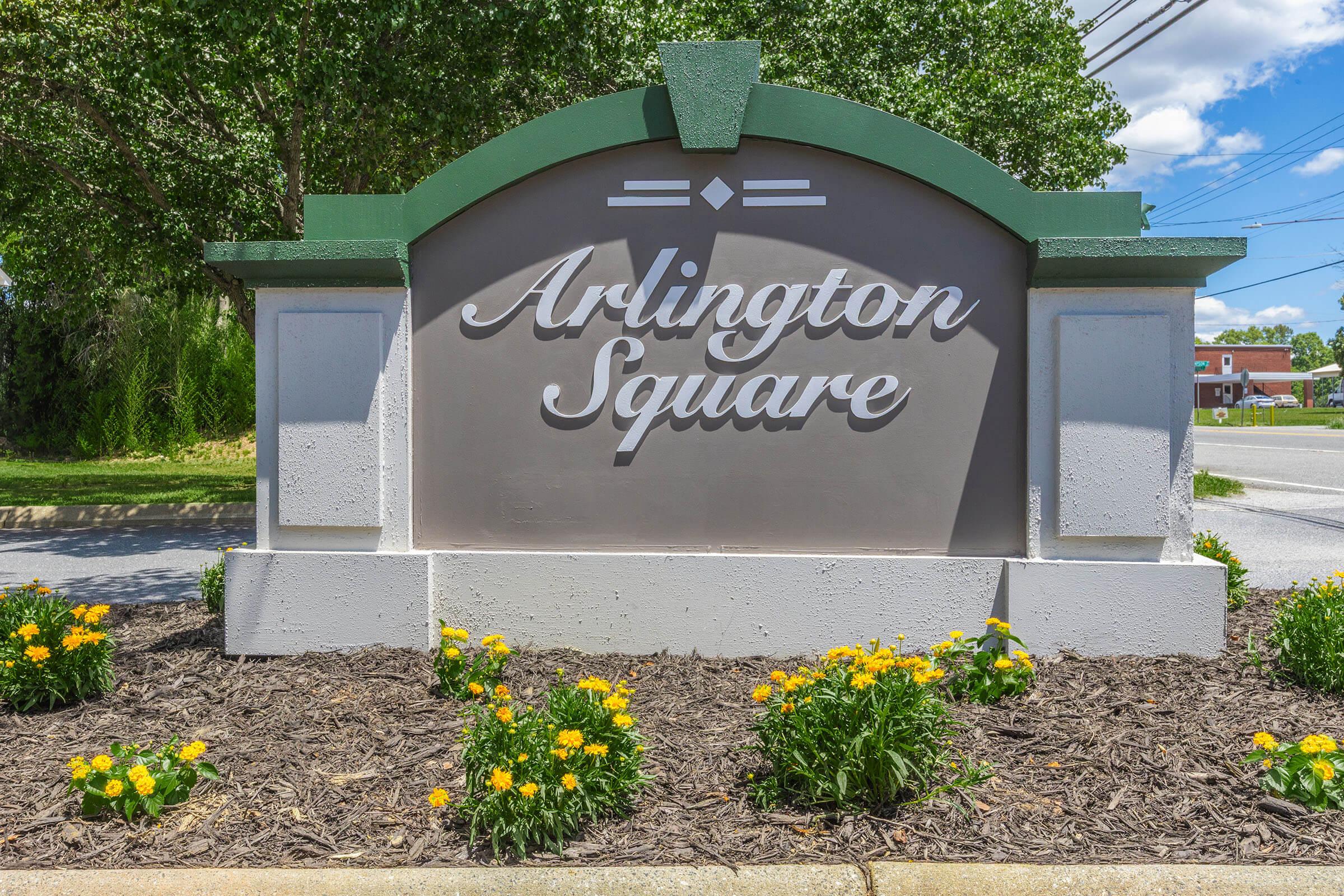 Sign for Arlington Square, featuring elegant lettering against a gray background, framed by green decorative elements. The sign is set in a landscaped area with yellow flowers and green plants, under a clear blue sky with some clouds in the background.