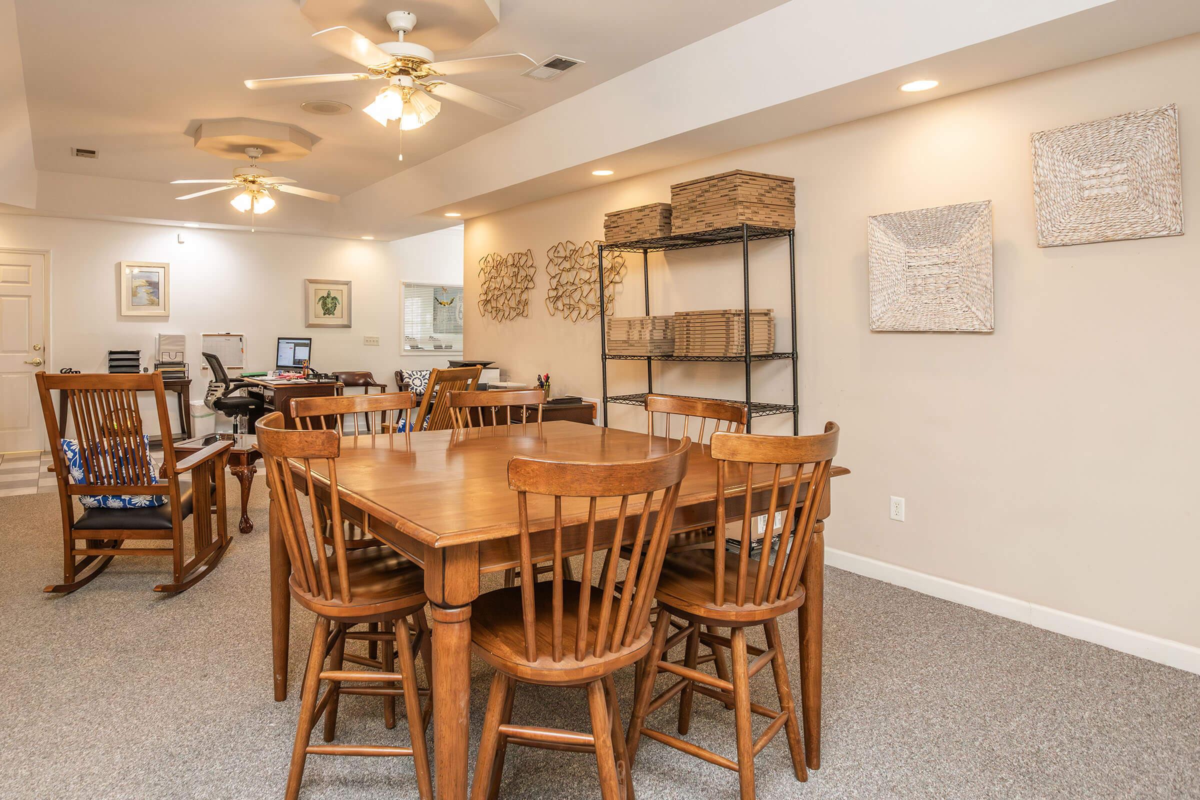 A well-lit dining area featuring a wooden table surrounded by chairs, with a decorative shelving unit in the background. The walls are adorned with artwork, and the space has a cozy, inviting atmosphere, ideal for gatherings or meals. Ceiling fans add to the comfort of the room.