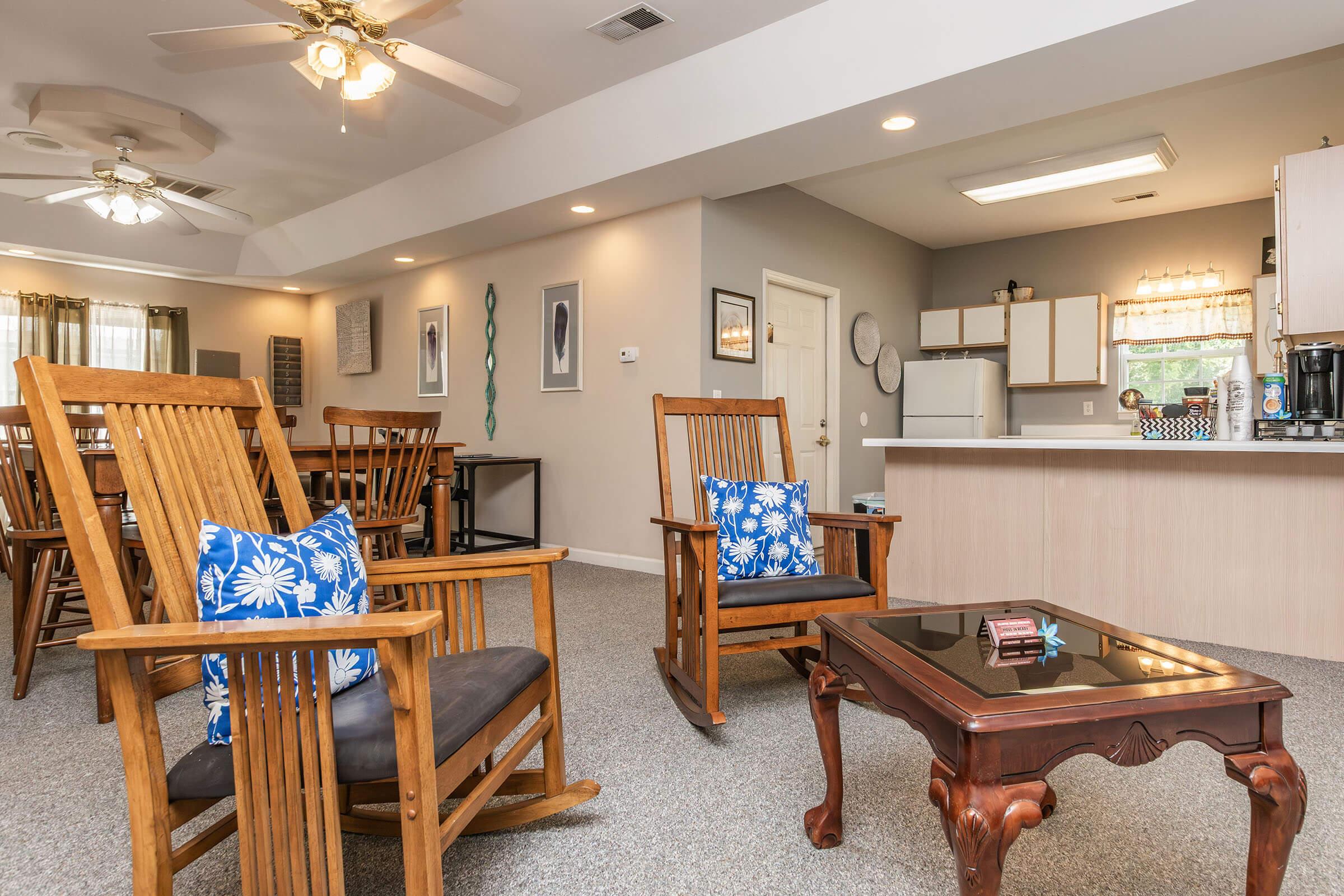 A cozy living space featuring two wooden rocking chairs with blue floral cushions, a dark wooden coffee table, and a kitchenette in the background. The room has a ceiling fan, warm lighting, and decor on the walls, creating an inviting atmosphere for relaxation and socializing.