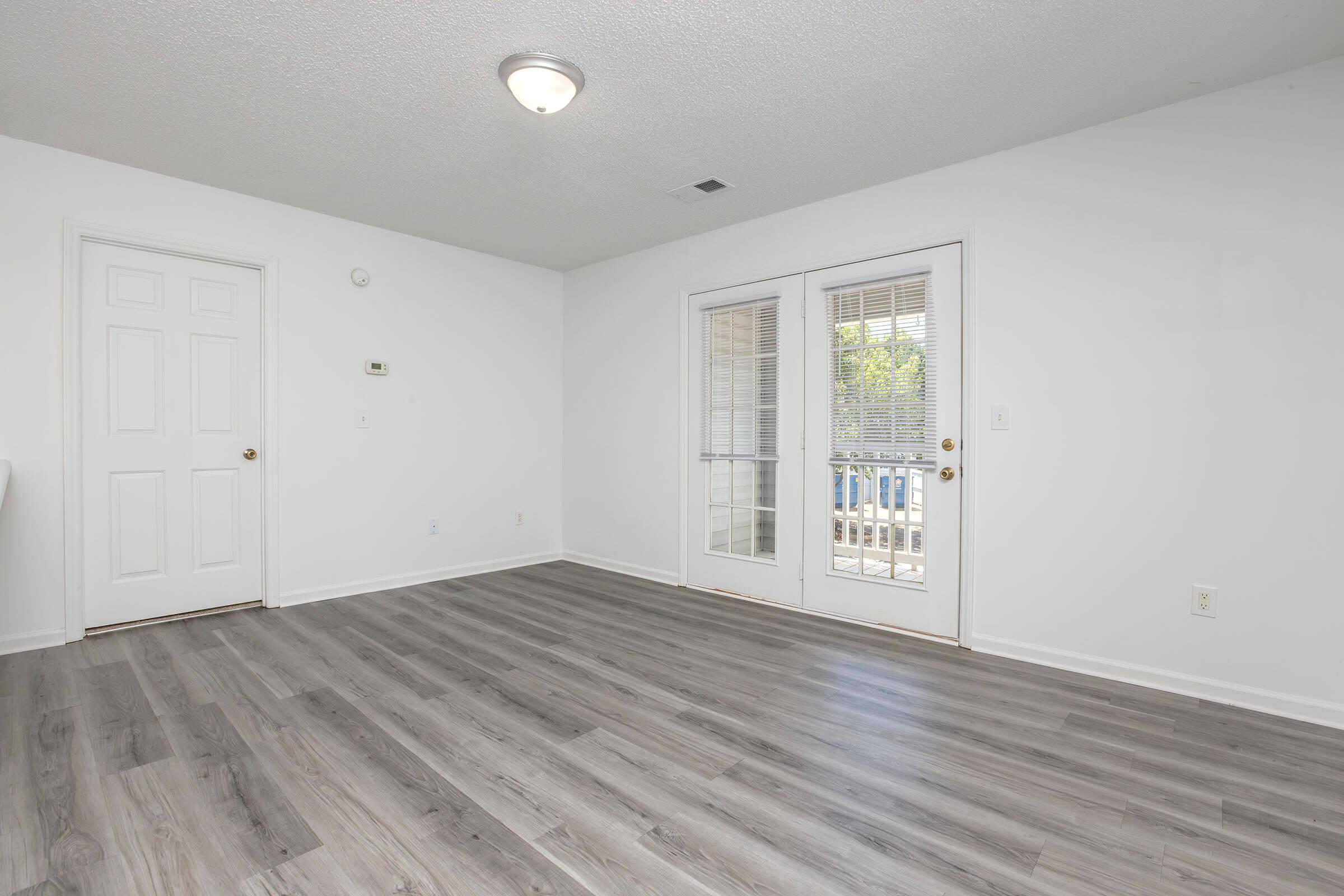 Empty room with light gray walls and a windowed door leading outside. The floor features light-colored laminate, and there's a doorway visible on the left. Natural light illuminates the space, creating a bright and open atmosphere.