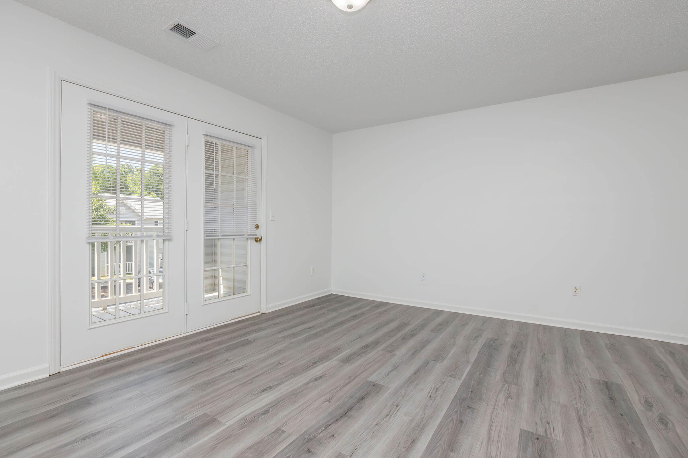 A spacious, empty room featuring light-colored walls and modern laminate flooring. It has a set of double doors that open onto a small balcony, letting in natural light. The room is devoid of furniture, highlighting its potential for personalization and decoration.