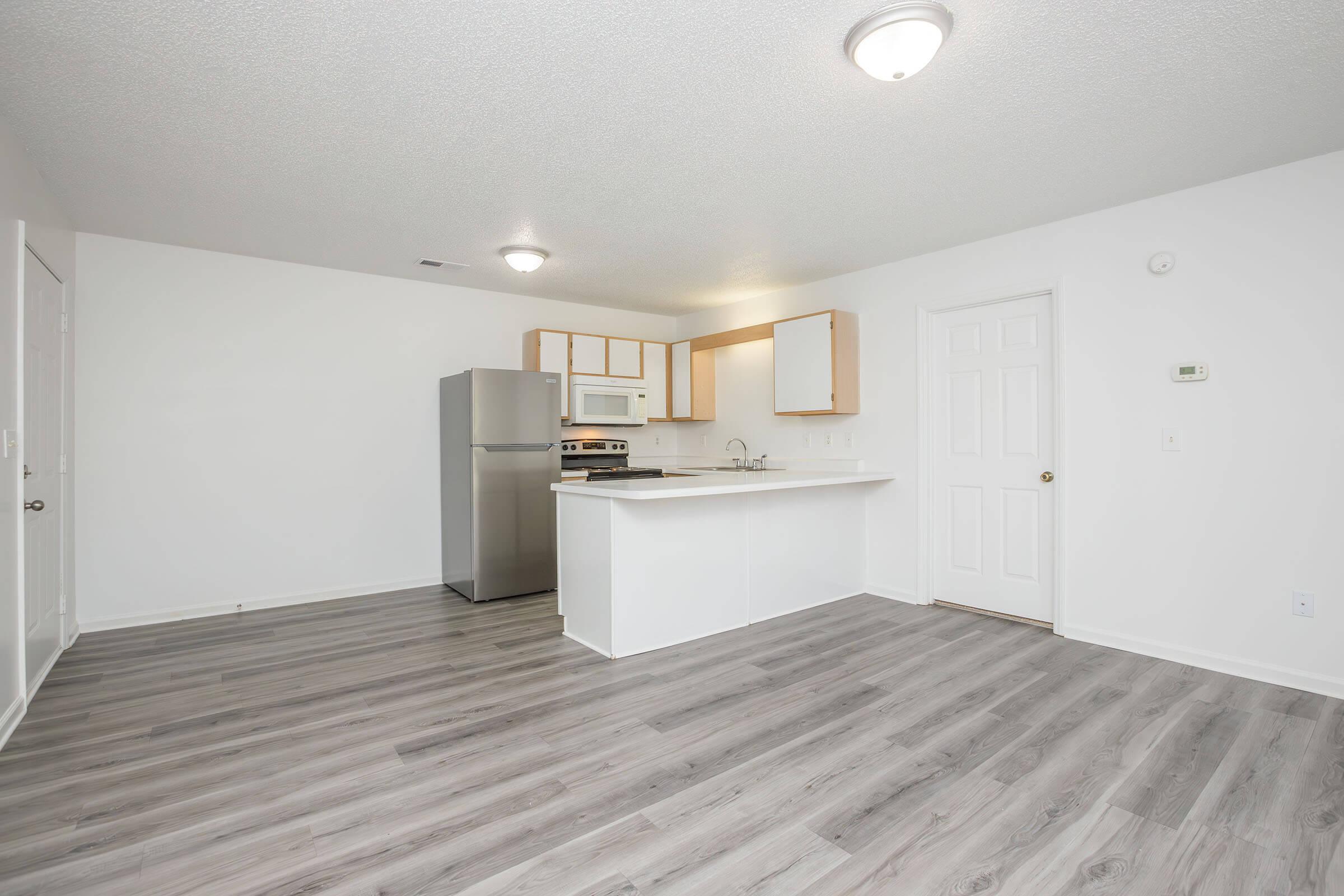 A view of a modern kitchen and living area featuring stainless steel appliances, a white countertop, and light wood cabinetry. The floor is covered in light gray laminate, and the walls are painted white. A door and a thermostat are visible, contributing to a clean and open layout.