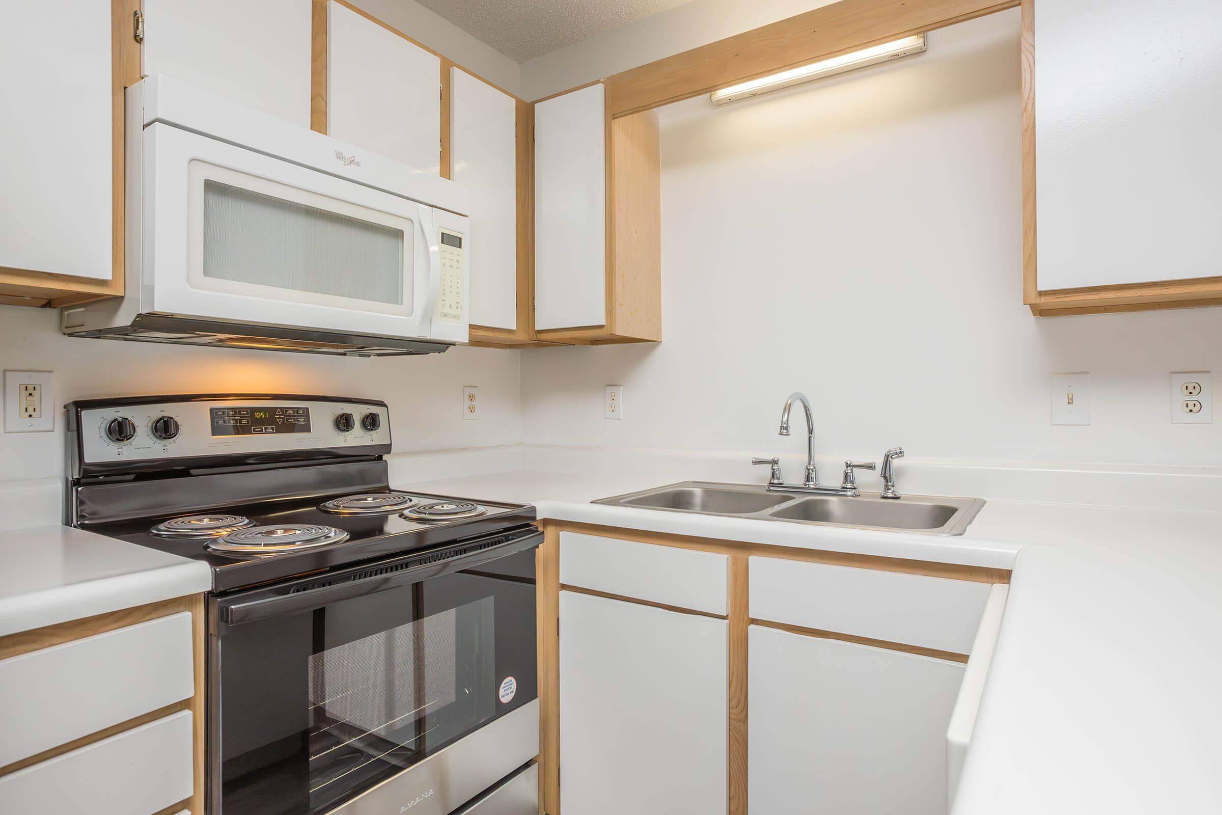 Modern kitchen with white countertops and cabinets. Features a black electric range with an oven, a microwave above it, and a double sink with a shiny faucet. The space is well-lit with under-cabinet lighting, offering a clean and functional atmosphere.