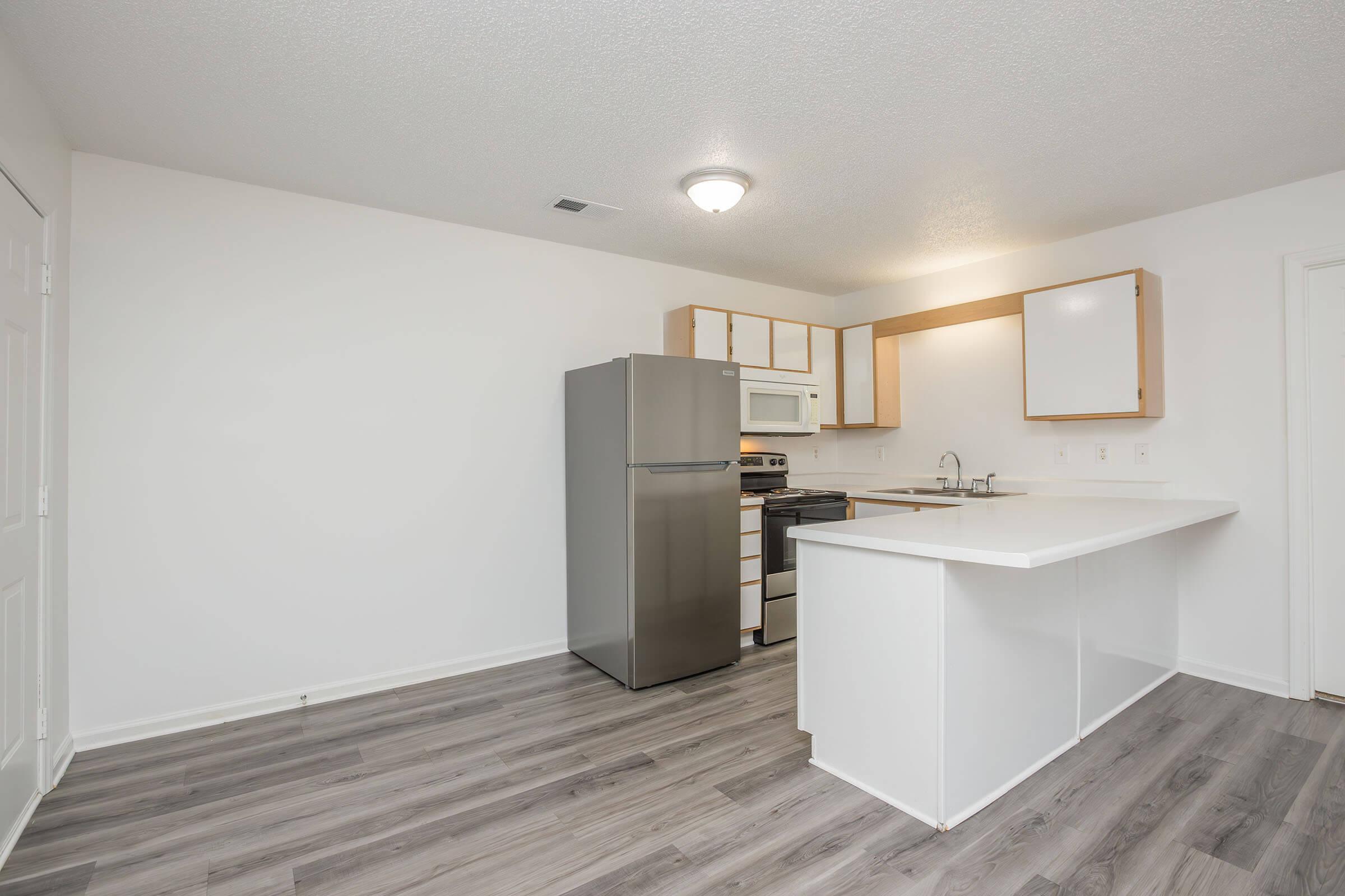 Modern kitchen featuring stainless steel refrigerator and microwave, white cabinetry, and a gas stove. The design includes a spacious island with a countertop and light wood flooring. The walls are painted white, creating a bright and airy atmosphere.