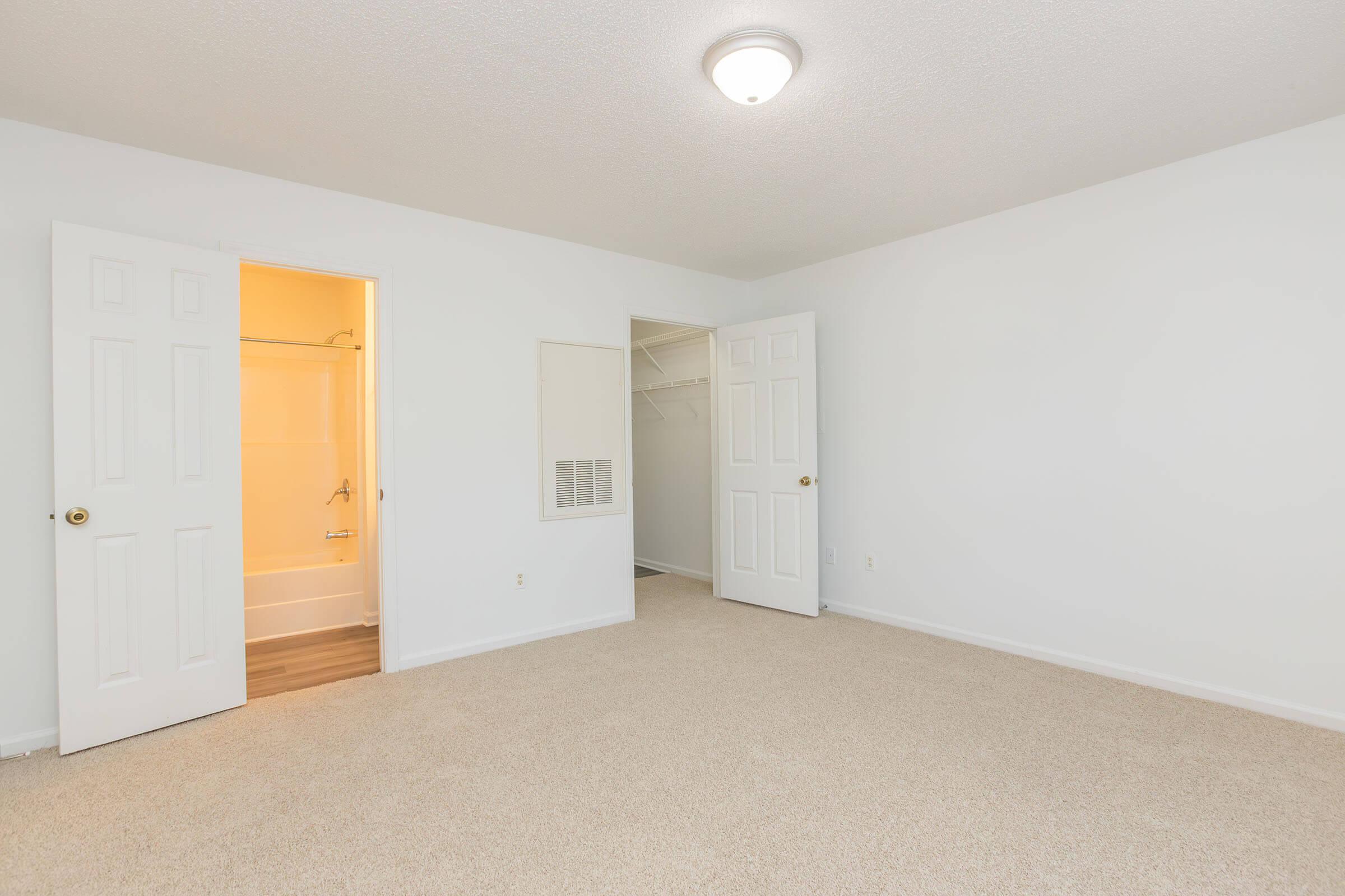 A spacious, empty room with beige carpet and white walls. Two open doors lead to a bathroom and a closet. The room is well-lit by a ceiling light fixture, creating a bright and inviting atmosphere.
