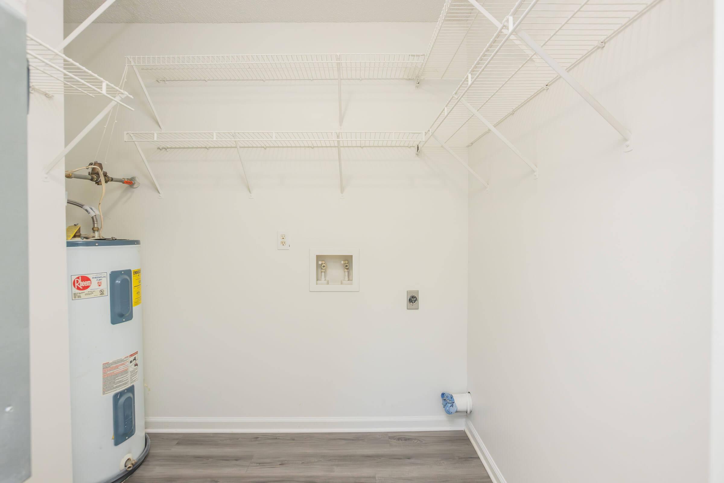 A spacious laundry room featuring white walls and light grey vinyl flooring. There's a water heater on the left side, and metal wire shelving mounted along the wall for storage. Two electrical outlets are visible, and the room is well-lit, creating an open and organized atmosphere.