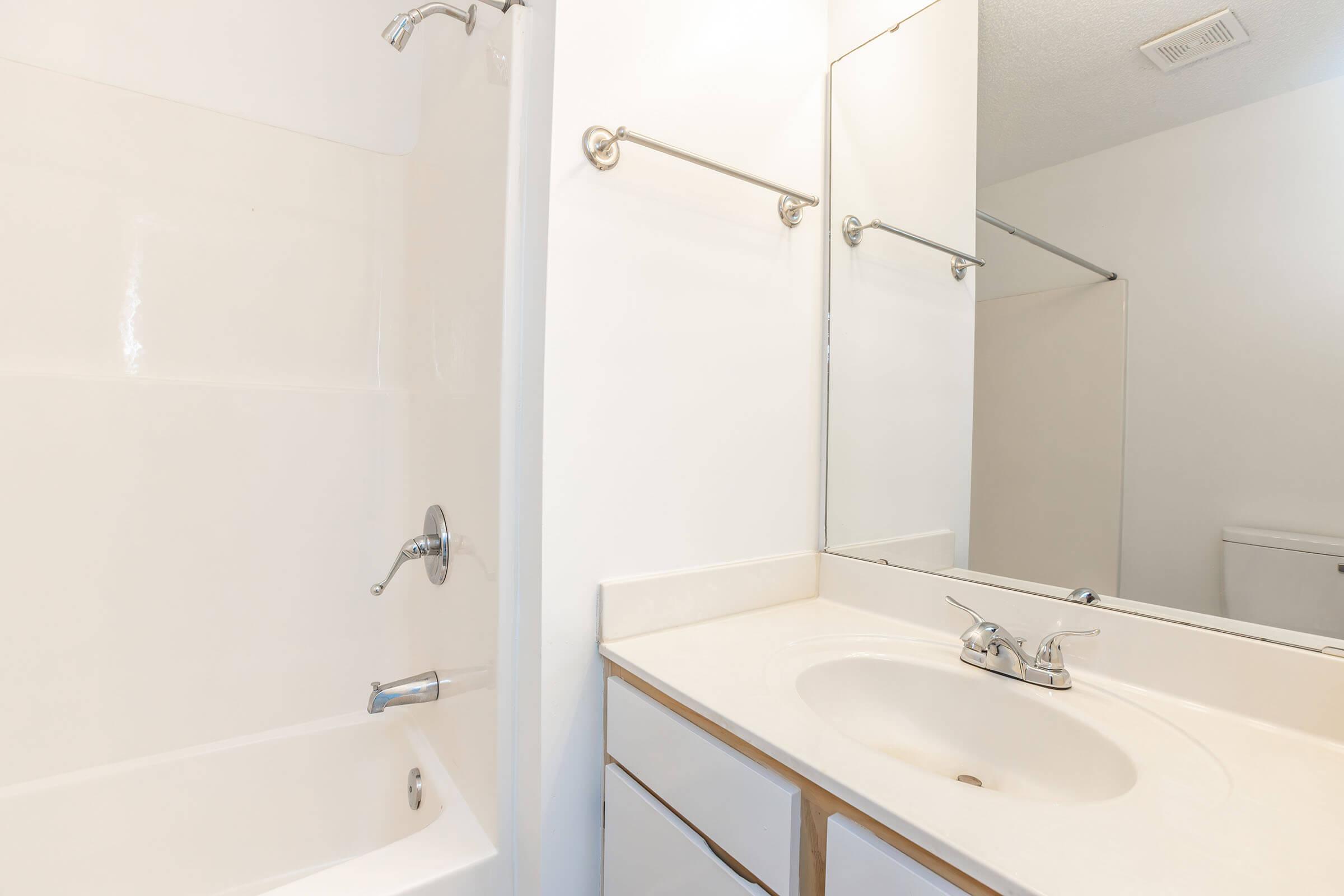 A clean, modern bathroom featuring a beige tub with a showerhead, a sink with a faucet, and a large mirror above the sink. The walls are painted white, and there are towel bars on the walls. In the background, a glimpse of a toilet can be seen.