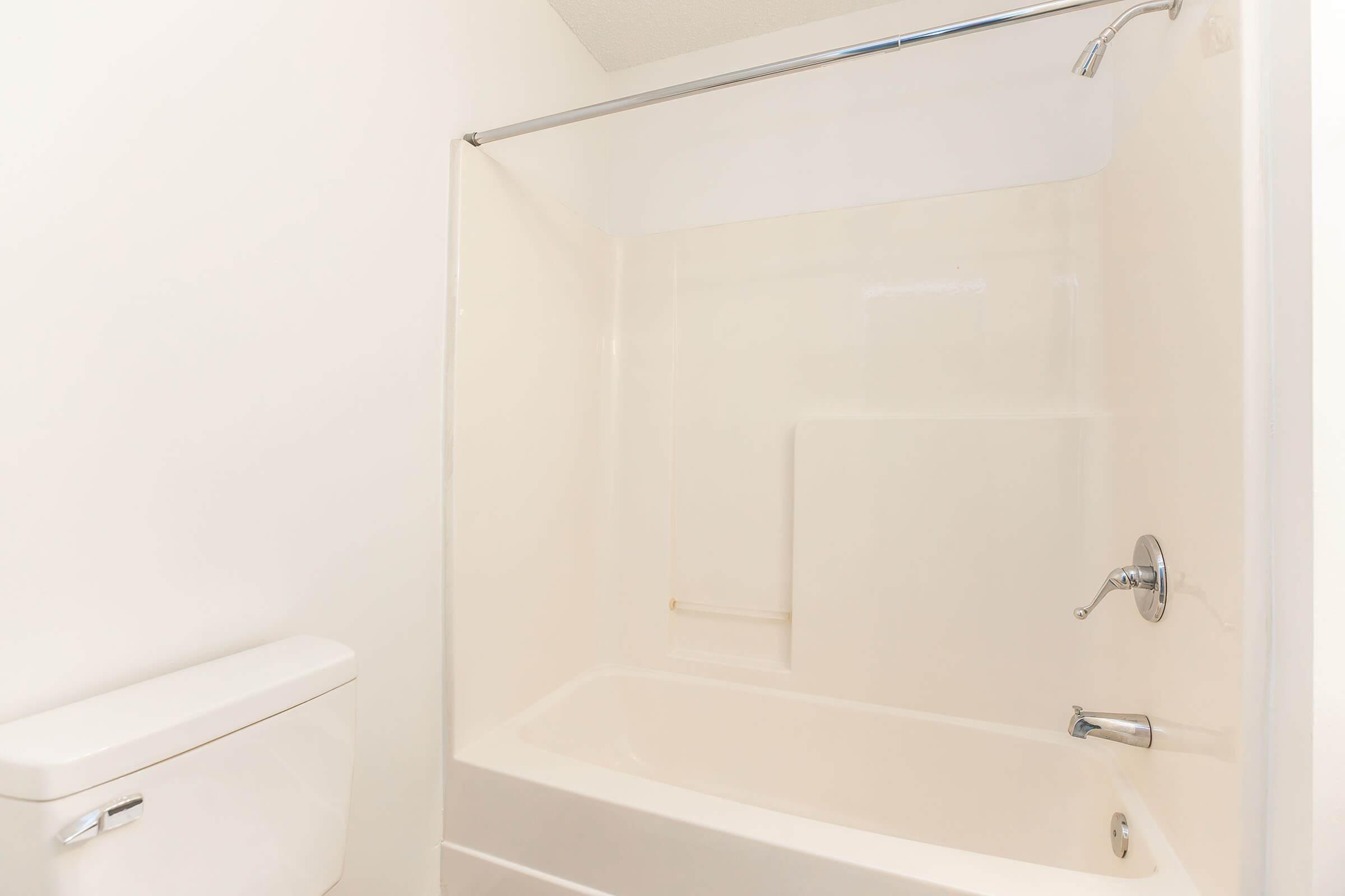 A clean, minimalistic bathroom featuring a white bathtub with a shower curtain rod, a showerhead, and a wall-mounted faucet. To the left, there is a white toilet. The walls and surfaces are light-colored, creating a bright and airy atmosphere.