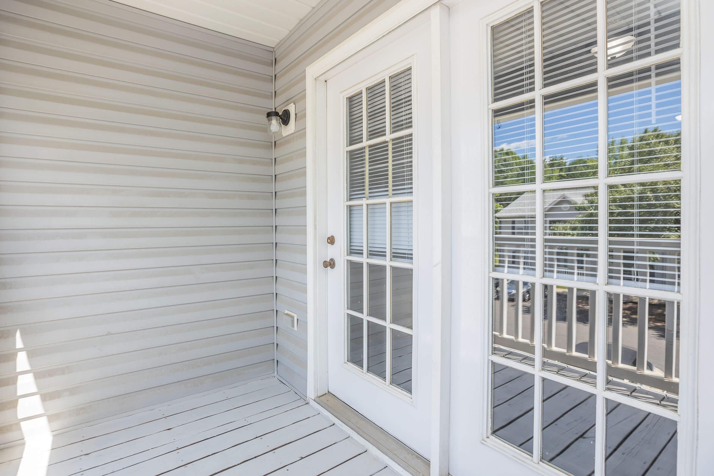 A well-lit entryway featuring a pair of white French doors with glass panels and blinds. The walls are light gray with horizontal siding, and the floor is made of wooden planks. Natural light floods the space, creating a welcoming atmosphere.