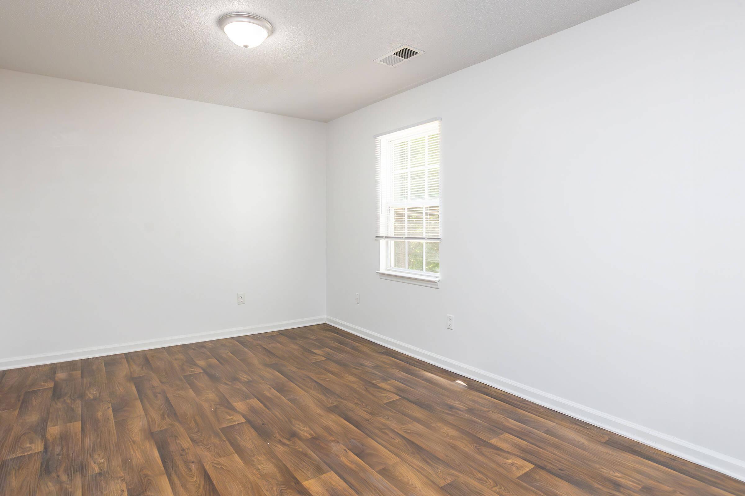 Empty room with light-colored walls and dark wood-like flooring. A single window lets in natural light, and a ceiling light fixture is present. The space appears well-lit and spacious, indicating a fresh, minimalist design.