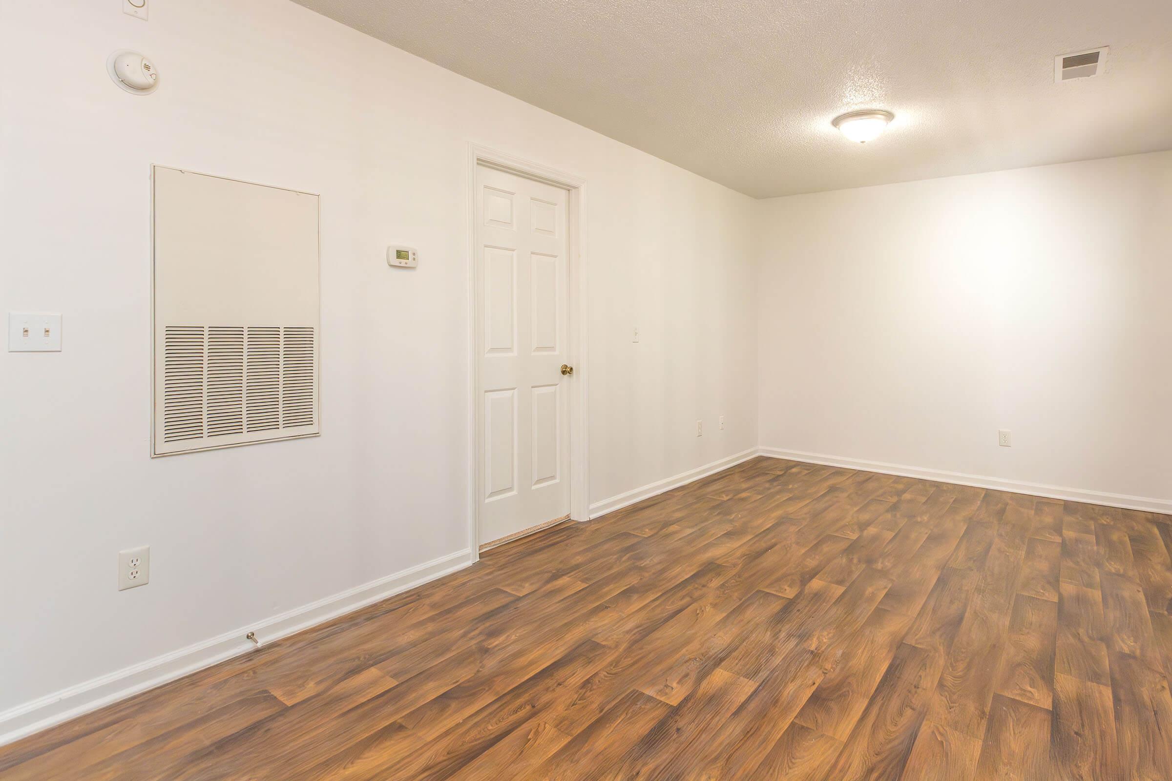 Empty room featuring light-colored walls, a wooden floor, a wall-mounted heater, a thermostat, and a closed door. Soft overhead lighting illuminates the space, creating a clean and open atmosphere.