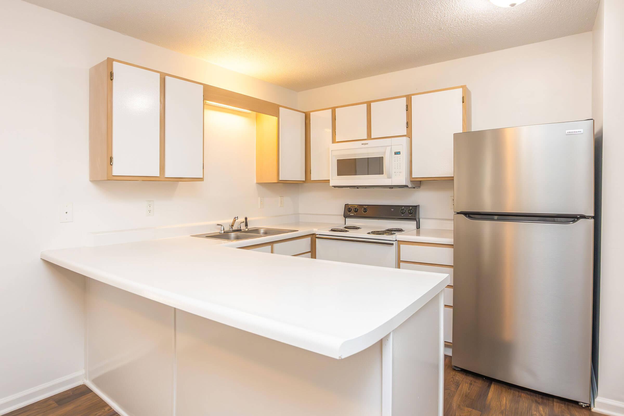 A clean, modern kitchen featuring white cabinetry with wooden accents, a stainless steel refrigerator, a microwave, and an electric stove. The countertop is spacious, with a double sink and a window above. The walls are painted a light color, enhancing the bright and airy feel of the space.