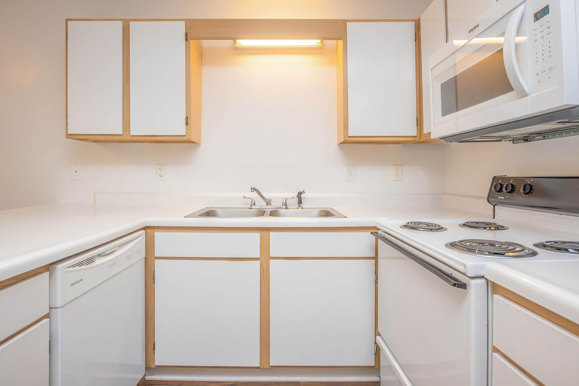 Modern kitchen with white cabinetry and countertops. Features a stainless steel sink, a built-in dishwasher, and an electric stove with four burners. A microwave is mounted above the stove, and a warm light is illuminating the workspace. The walls are painted in a neutral color, creating a bright and clean atmosphere.