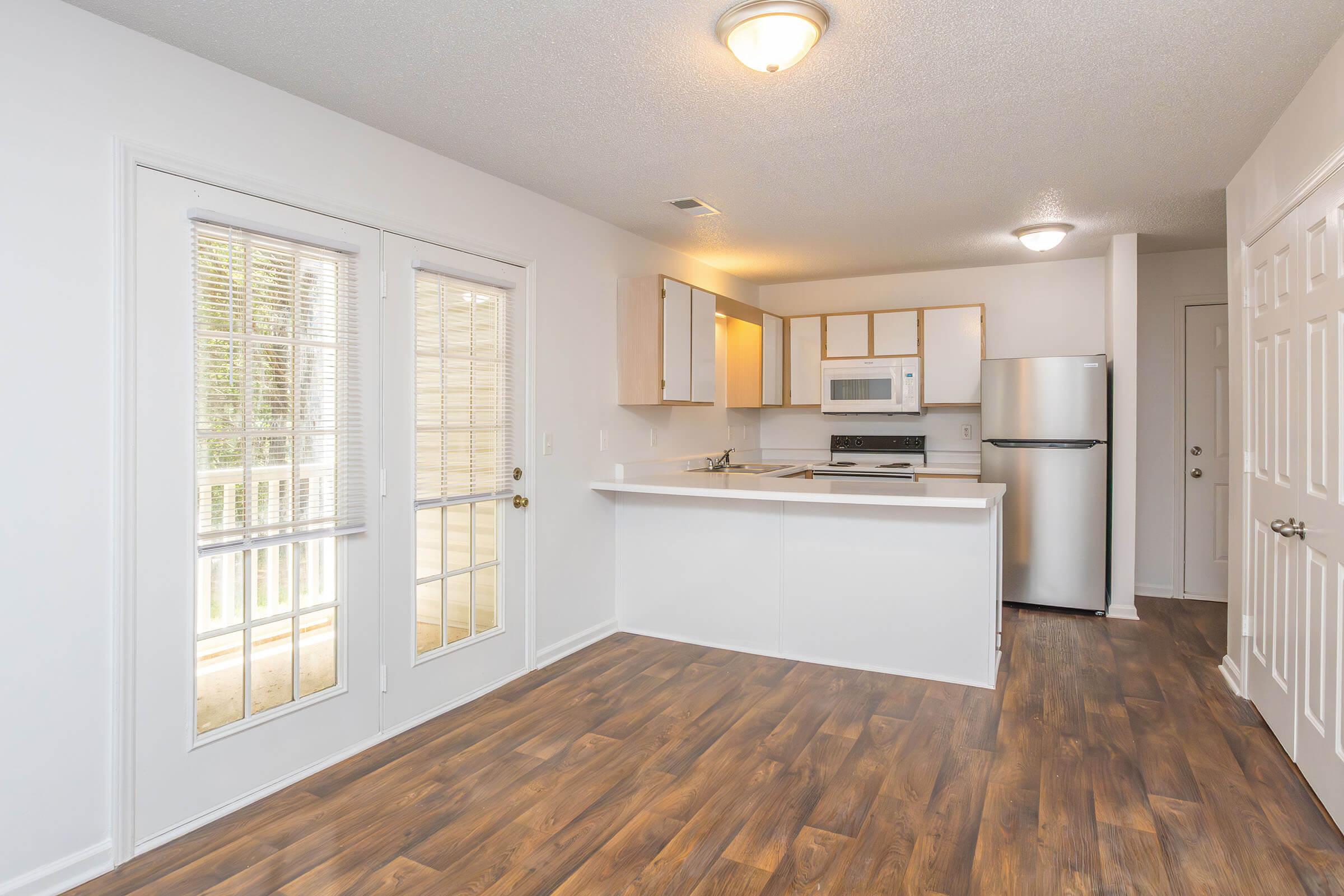 Bright and modern kitchen with light wooden cabinets, a white countertop, and stainless steel appliances. The space features sliding glass doors that lead to an outdoor area, and warm-toned laminate flooring adds a cozy touch. The room is well-lit, showcasing a clean and inviting atmosphere.