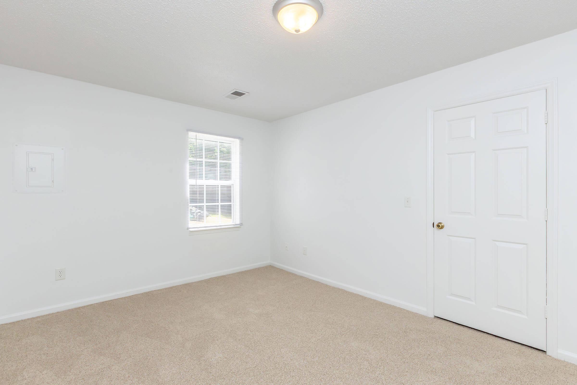 A vacant room with white walls and beige carpet. A single window allows natural light to enter, while a light fixture is mounted on the ceiling. There is a closet door on the right and a blank wall with an electrical panel on the left. The space appears clean and ready for furnishing.