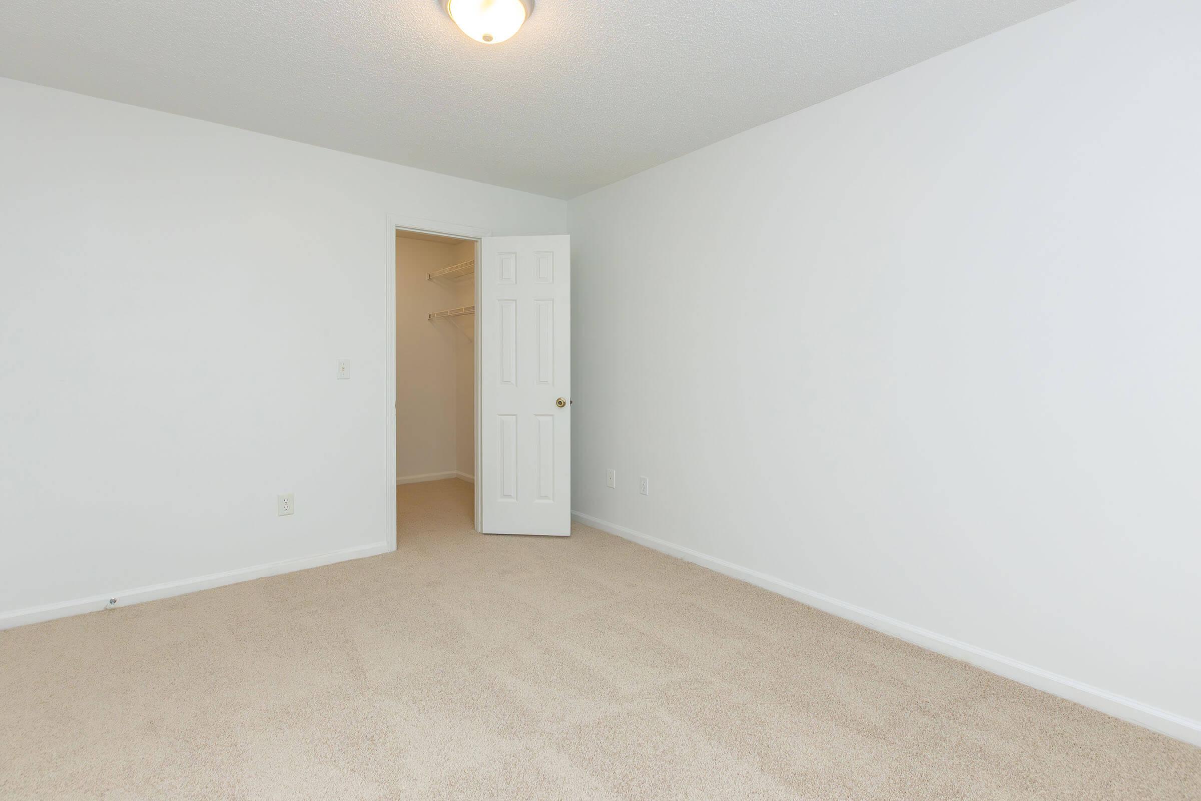 A clean, empty bedroom with beige carpeting, white walls, and a ceiling light. A closet with an open door is visible on the left, showcasing shelving inside. The room has a bright and airy feel, suitable for personal decoration or furniture arrangement.