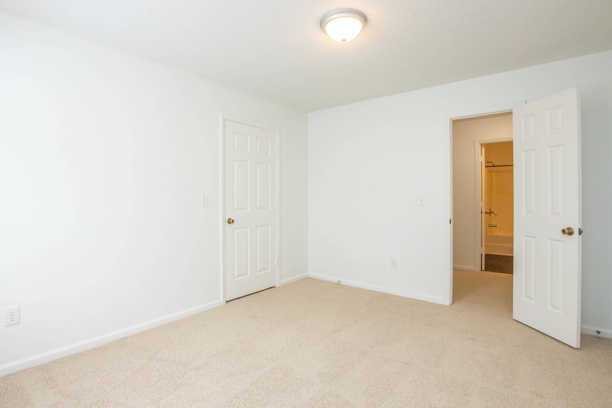 A vacant, unfurnished room with beige carpet and white walls. The room features a ceiling light and an open door leading to a bathroom. Another closed door is on the left side of the image, indicating a closet or storage space. The overall atmosphere is bright and minimalistic.