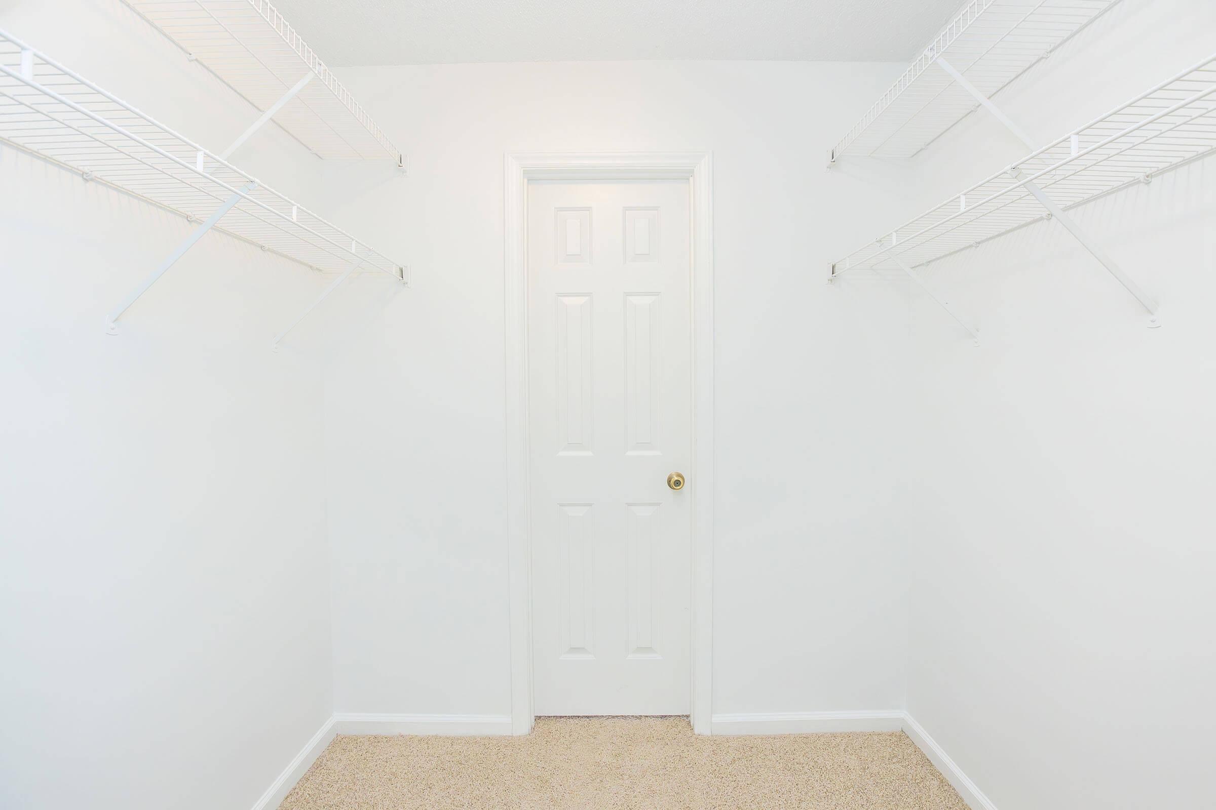 Empty closet with white walls and a closed white door. There are two wire shelving units mounted on the walls, one on each side. The floor is carpeted in a neutral beige color, and there is ample natural light illuminating the space.