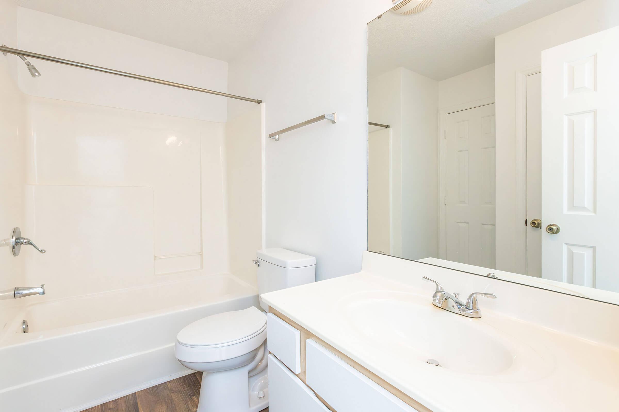 A clean, modern bathroom featuring a bathtub with a shower rod, a white toilet, and a sink with a mirror. The walls are painted white, and the floor has brown vinyl. A door can be seen in the background, suggesting a spacious layout.
