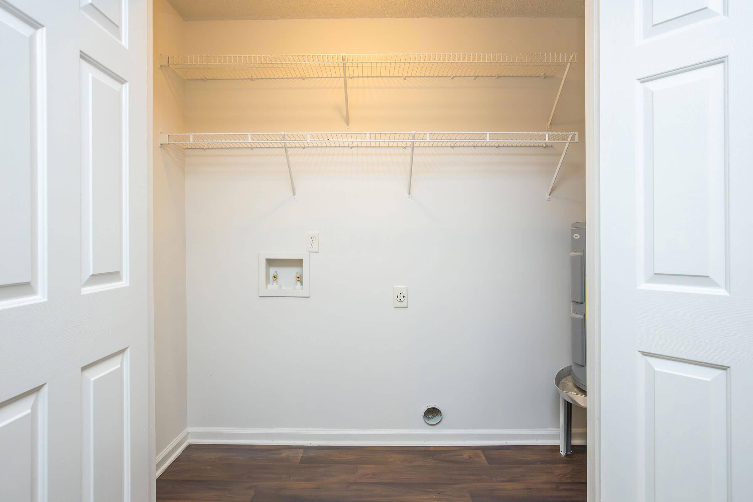 An empty closet space with white walls, featuring two wire shelves mounted on the back wall. There are two doors partially open, revealing the interior. A small electrical outlet and a water connection are visible on the wall. The flooring is a dark wood pattern, adding contrast to the light color scheme.