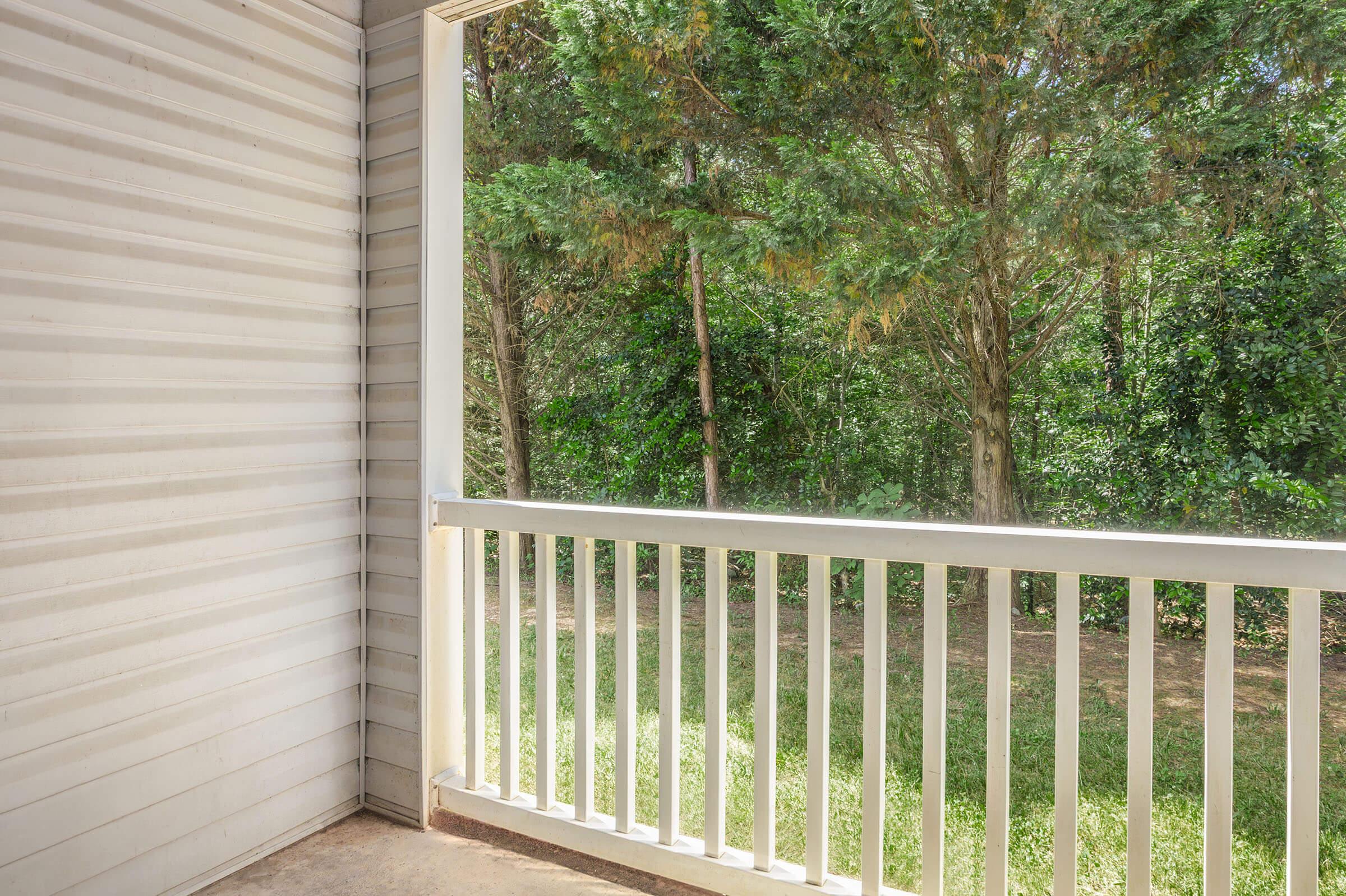 A spacious balcony with a white railing, overlooking a lush green area filled with trees and shrubs. Natural light filters in, creating a serene outdoor space perfect for relaxation.