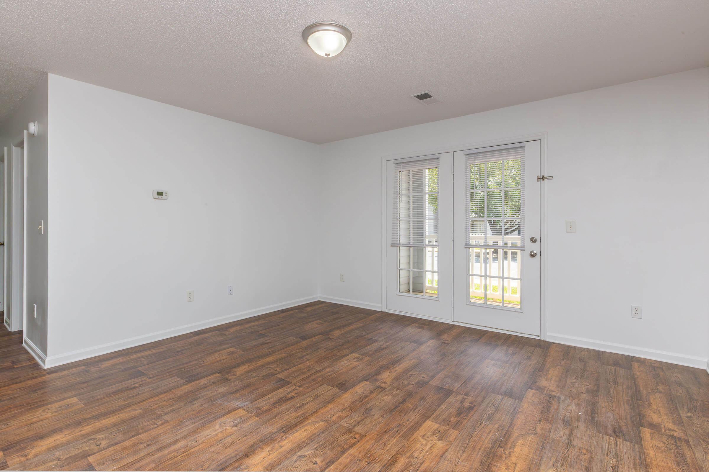 An empty room with light wood laminate flooring, white walls, and a ceiling light fixture. A set of glass doors leads to a small outdoor area, allowing natural light to enter. The room is spacious and uncluttered, suggesting it could be used for various purposes.