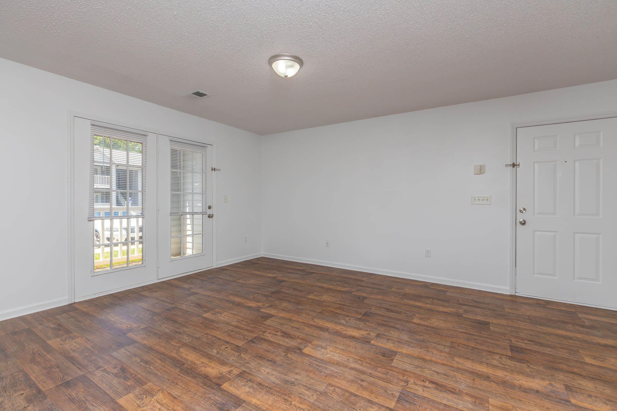 A spacious, empty room with light-colored walls and a wooden floor. Two windows provide natural light, and there are double doors leading outside. The room features a ceiling light fixture and is free of furniture, offering a blank canvas for decoration.