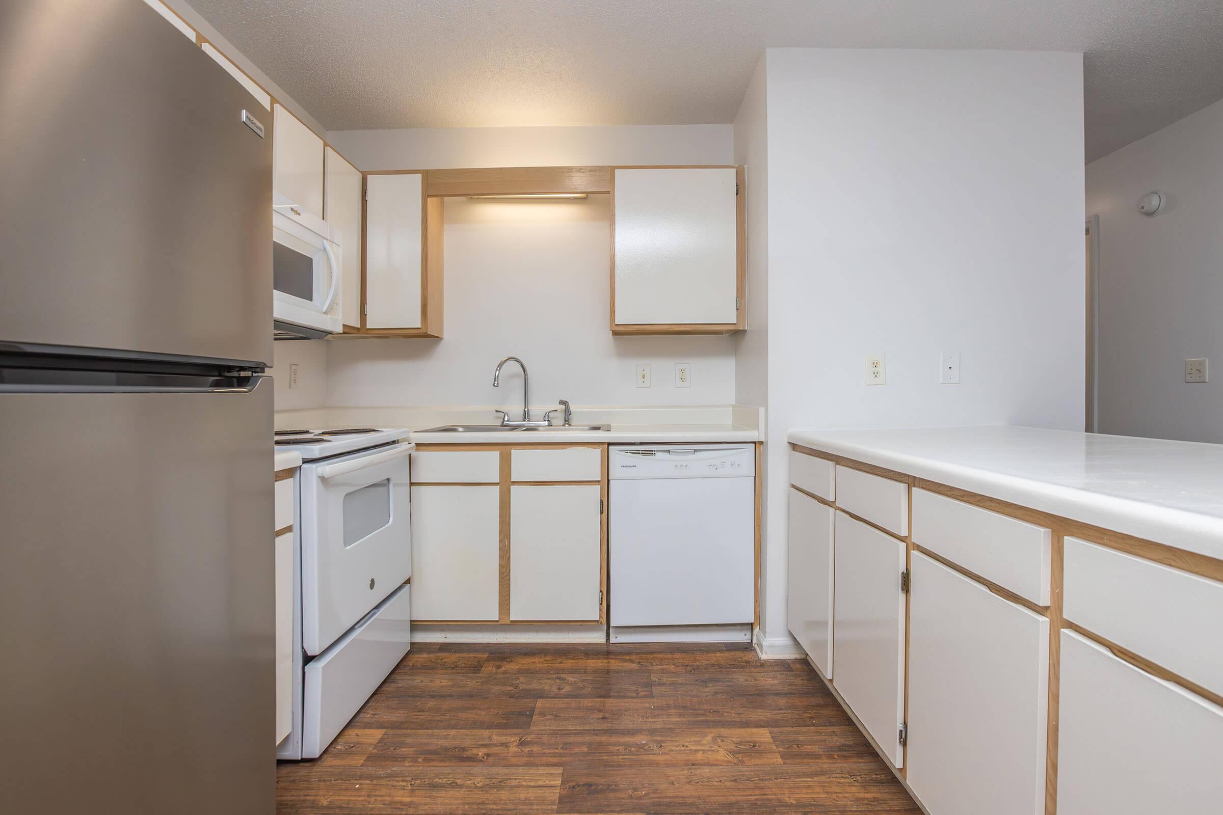 A modern kitchen featuring light-colored cabinets, a stainless steel refrigerator, and a microwave. The layout includes a sink, a dishwasher, and a stove, all with white appliances on a dark wood floor, creating a bright and spacious ambiance.