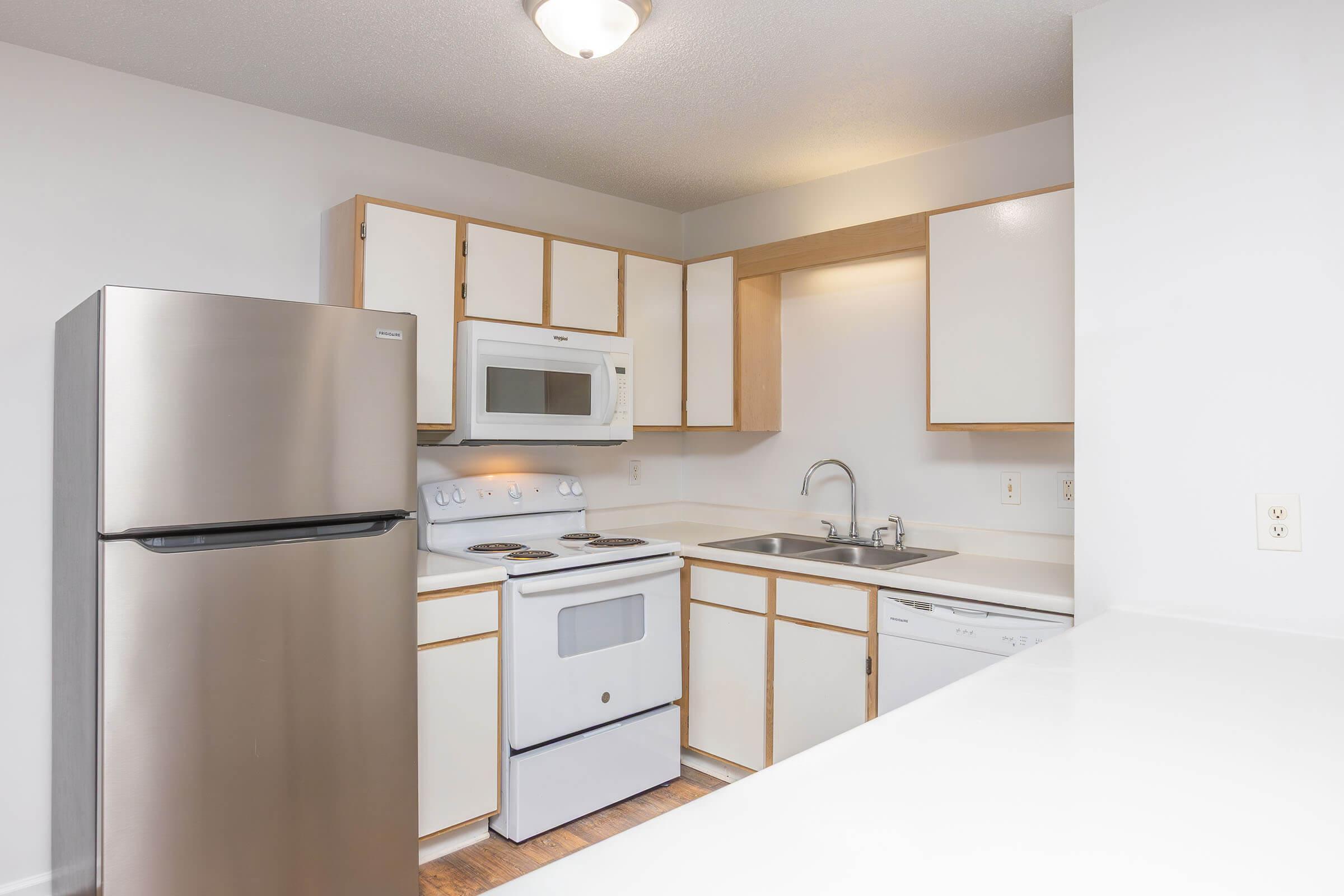 A modern kitchen featuring a stainless steel refrigerator, white microwave, stove, and dishwasher. The cabinets are light-colored with a simple design, and the countertops are white. Natural light is available, creating a bright and clean atmosphere. The floor is made of wood laminate.