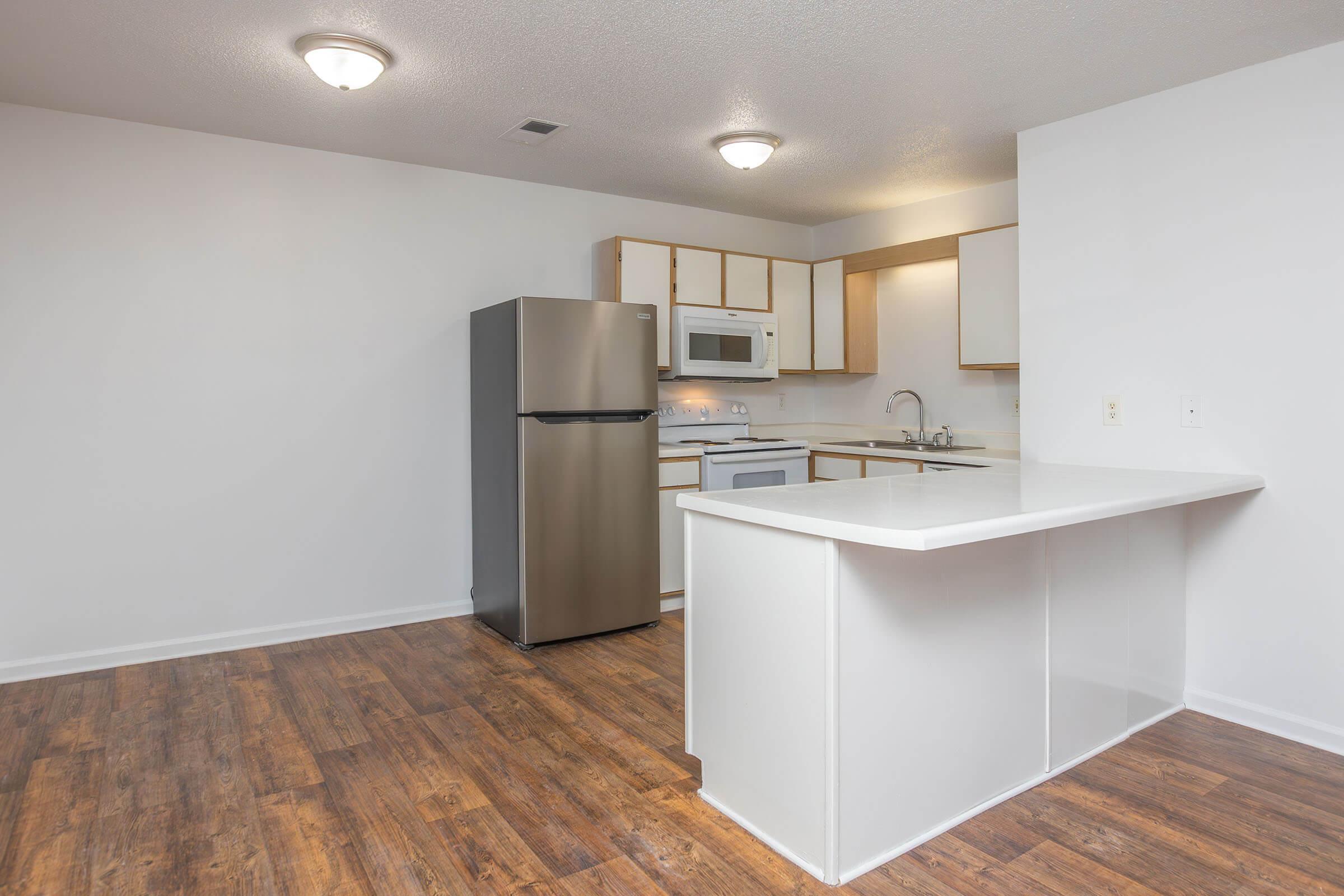 A modern kitchen in an apartment featuring a stainless steel refrigerator, white cabinetry, a countertop bar, and hardwood-style flooring. The space is well-lit with overhead lighting and has a clean, minimalist design.