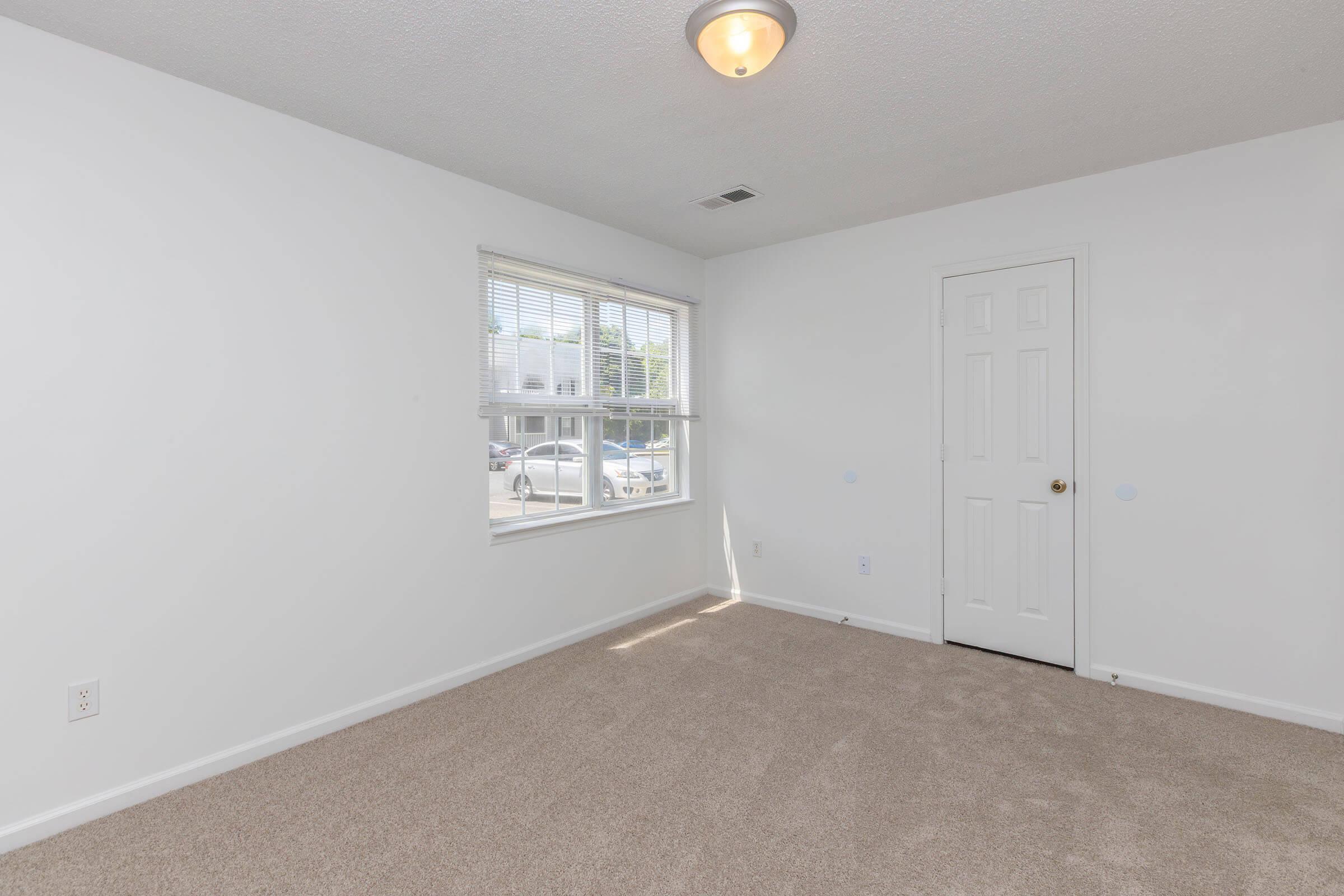 A bright, empty bedroom featuring light-colored walls, a carpeted floor, a window with blinds, and a door. Natural light enters through the window, illuminating the space. The room is unfurnished, creating a clean and open atmosphere, suitable for personalization.