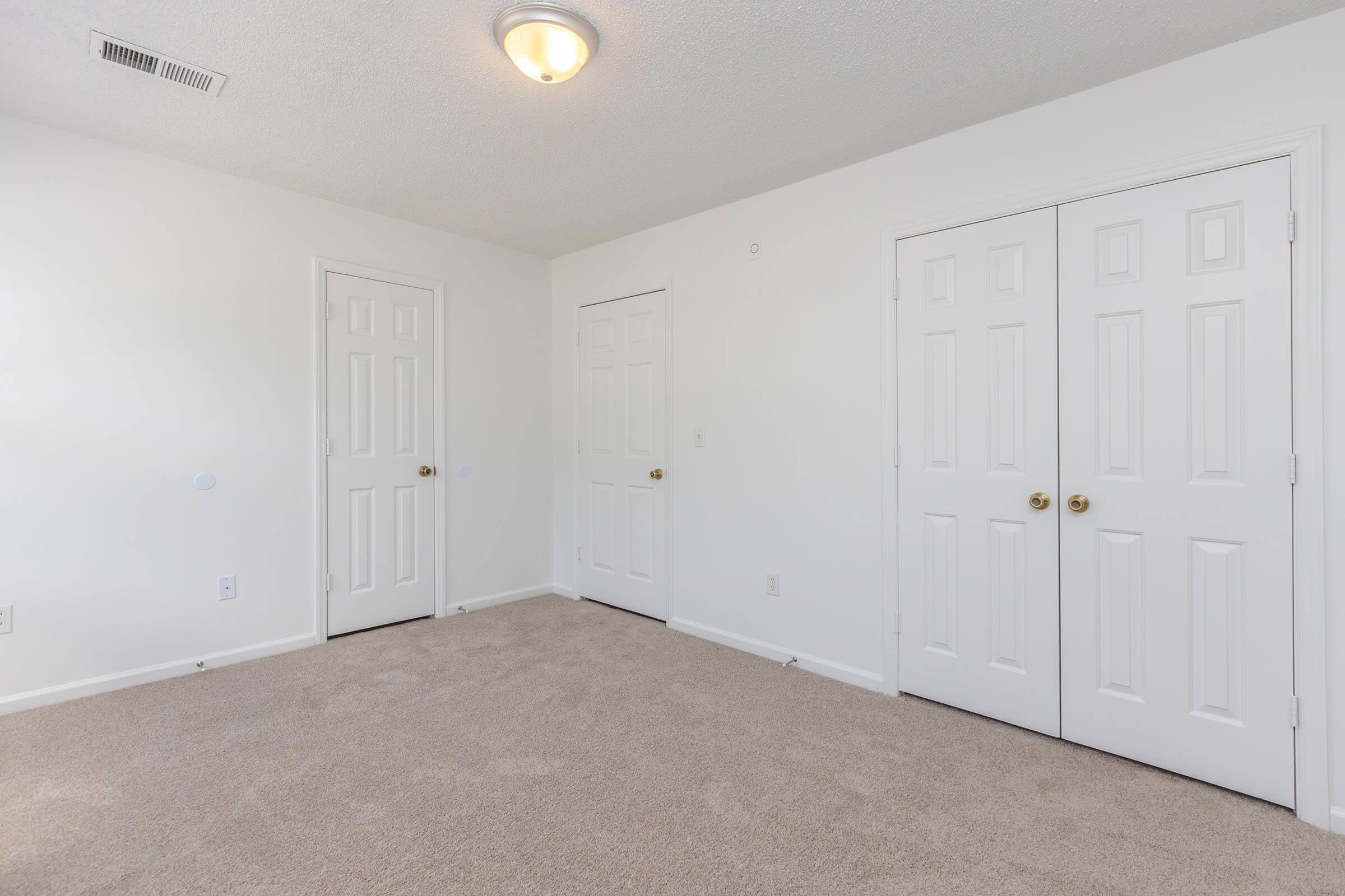 An empty, well-lit room with beige carpet, featuring white walls and two closed doors on the right. There is a ceiling light fixture in the center. The space appears tidy and unfinished, suitable for various furniture arrangements.