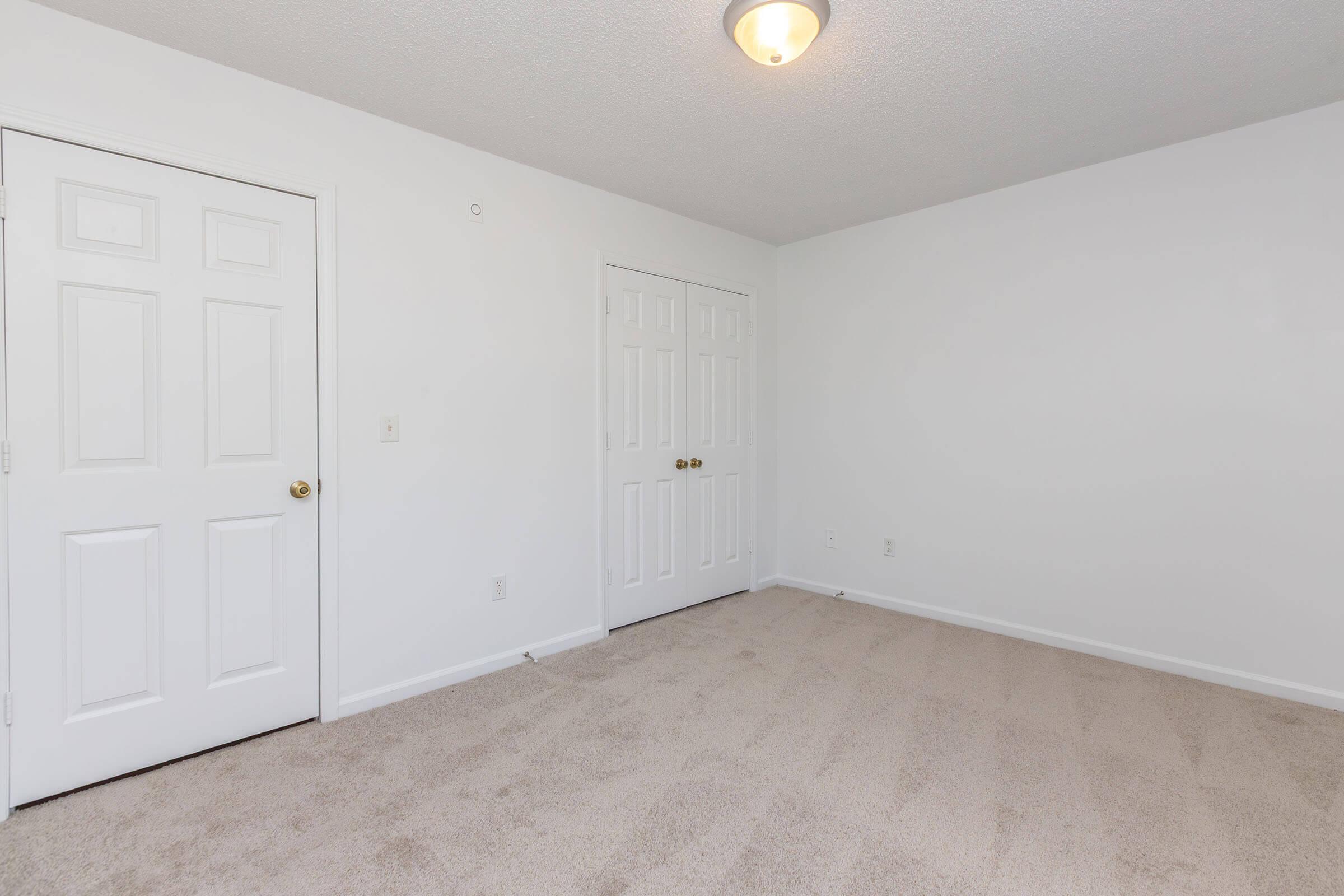 A large, empty room with light beige carpet and white walls. There are two closed doors against one wall, and the ceiling is illuminated by a central light fixture. The room appears clean and uncluttered, suitable for various uses like a bedroom or office space.