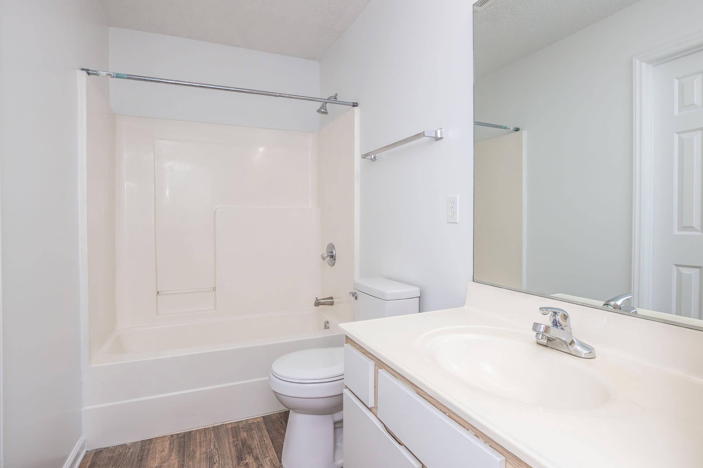 A clean, modern bathroom featuring a white bathtub with a sliding glass shower door, a toilet, and a single sink vanity with a mirror. The walls are painted light grey, and the floor has a wood-like texture. Natural light may be coming from a nearby window.