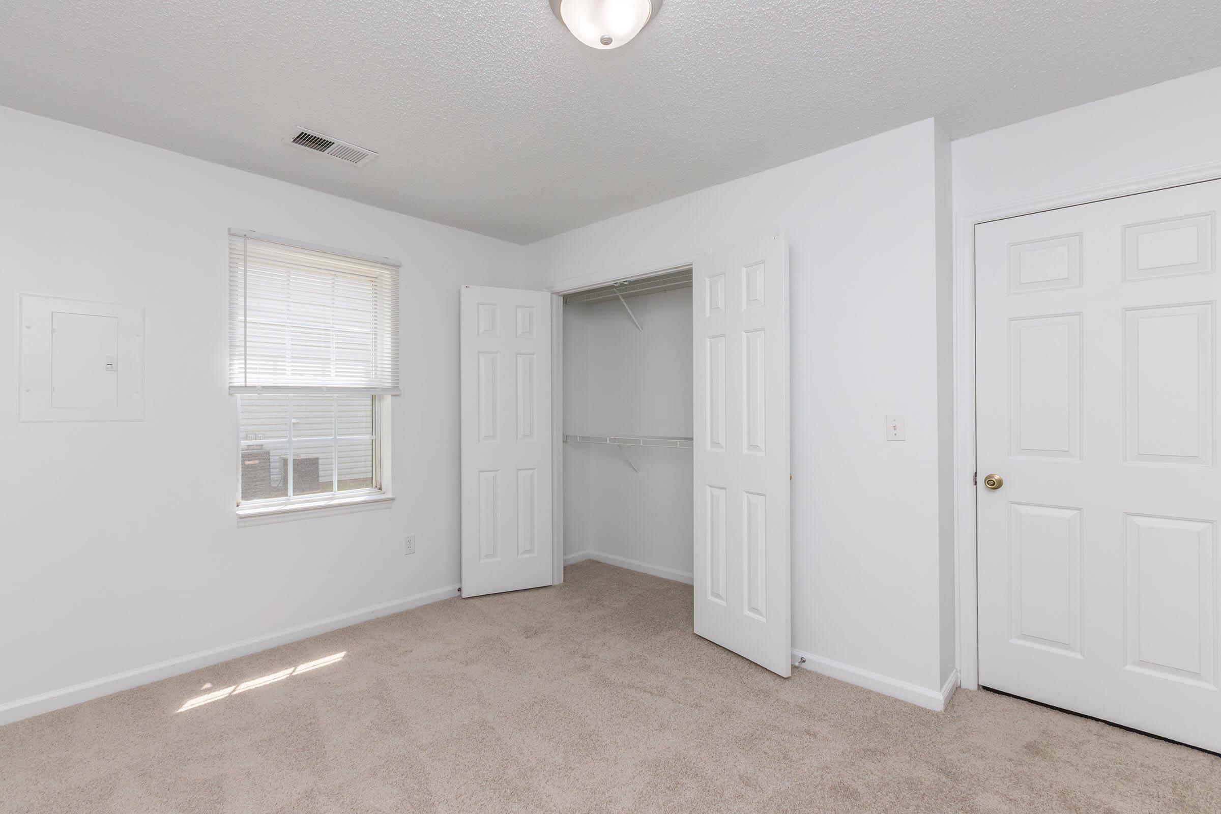 A bright and clean bedroom featuring beige carpet, white walls, and a ceiling light fixture. The room has two doors, one leading to a closet with sliding doors and another to a hallway. A window lets in natural light, and there is a small electrical panel visible on the left wall.