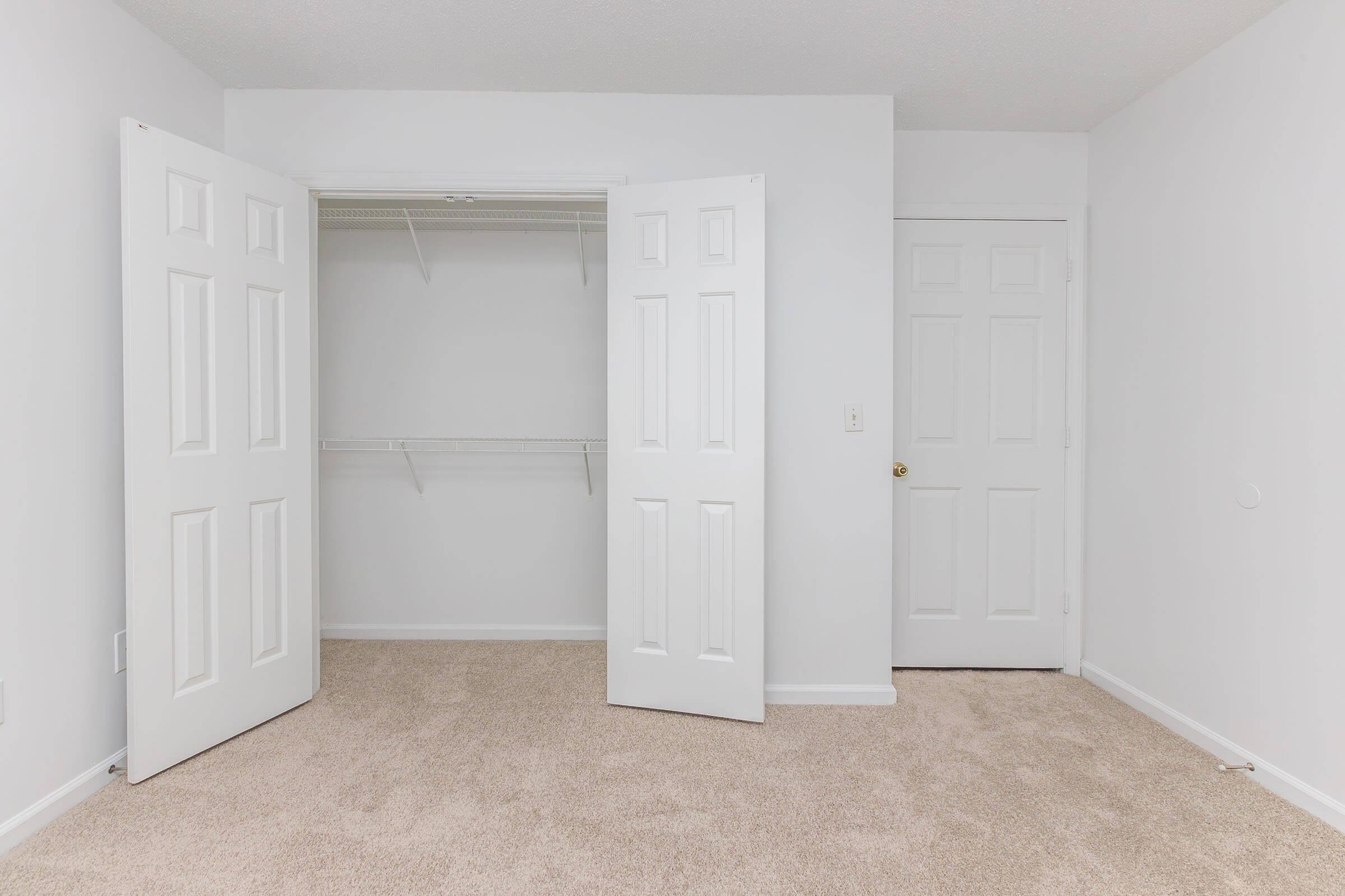 A spacious, empty room with light beige carpet. Two white closet doors are open, revealing a closet with a shelf but no items inside. A closed white door is visible on the right side, and the walls are painted white, giving the room a clean and bright appearance.