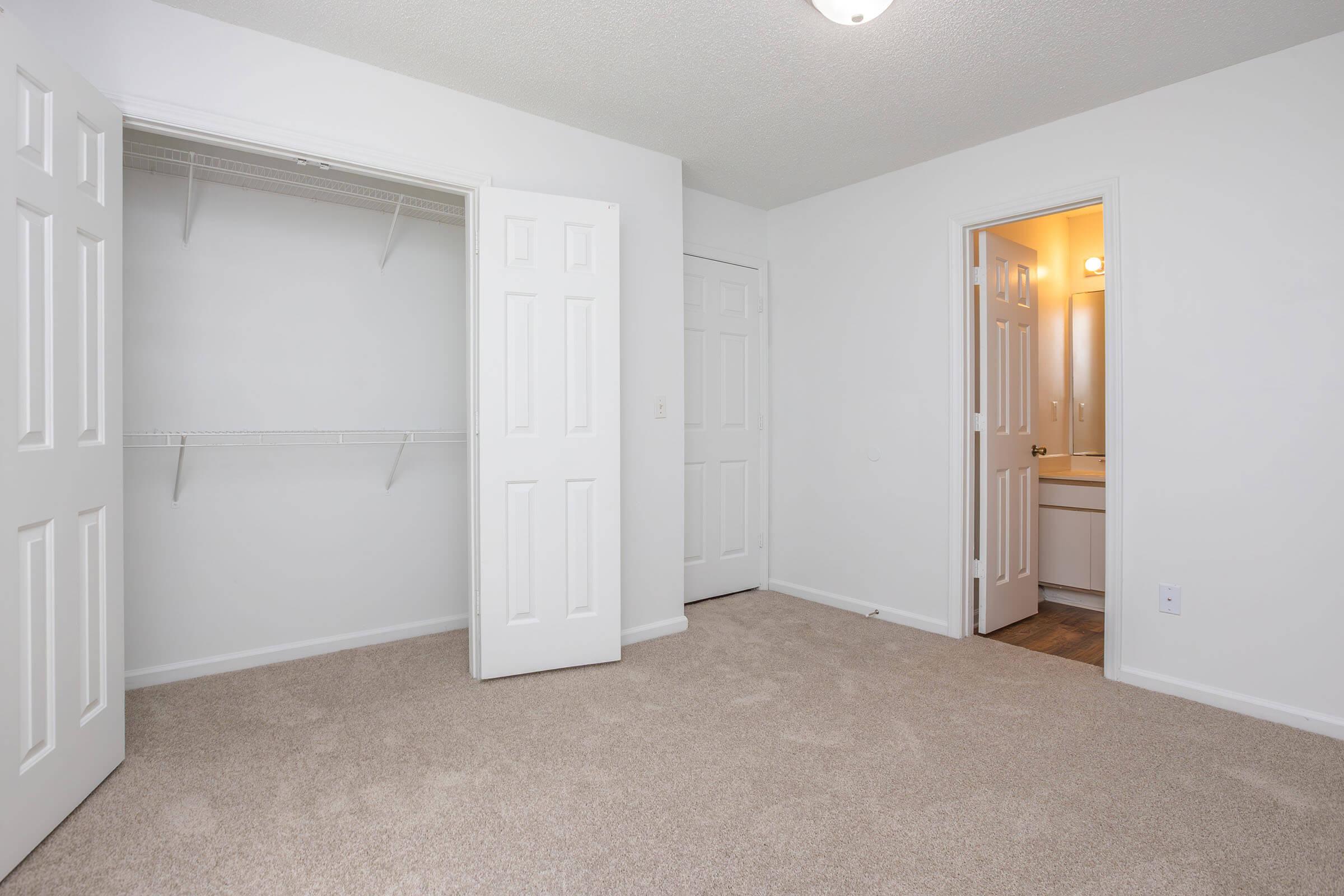 A clean, empty room with light beige carpet. Two white doors lead to a spacious closet and a bathroom. The walls are painted white, and a soft ceiling light is on. The room has a neutral, bright ambiance, making it feel open and airy.