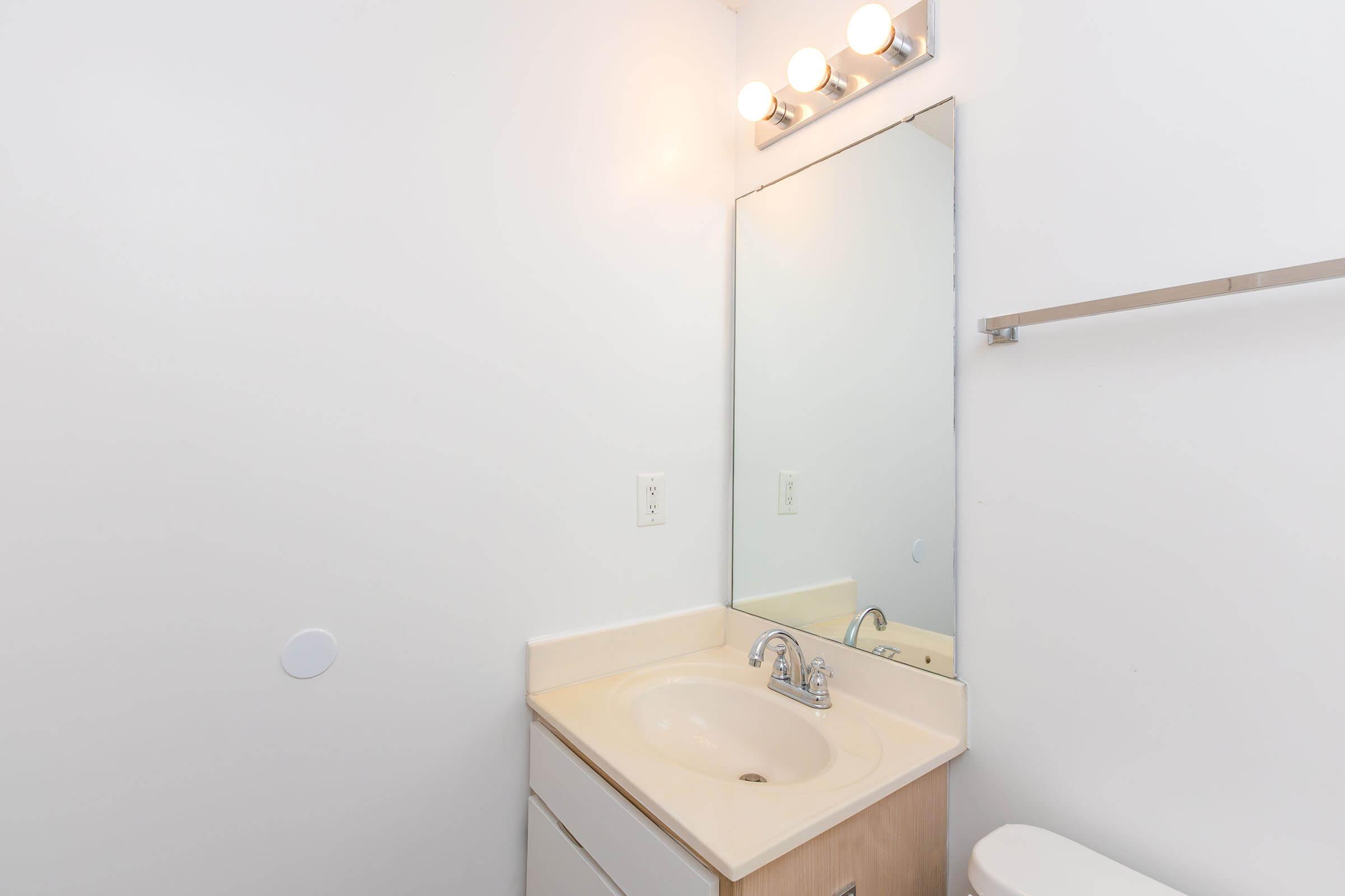 A clean and simple bathroom with a beige countertop sink, a large rectangular mirror above it, and a light fixture with three bulbs. The walls are painted white, and there's a towel bar mounted on the wall beside the mirror. The overall design is minimalistic and bright.