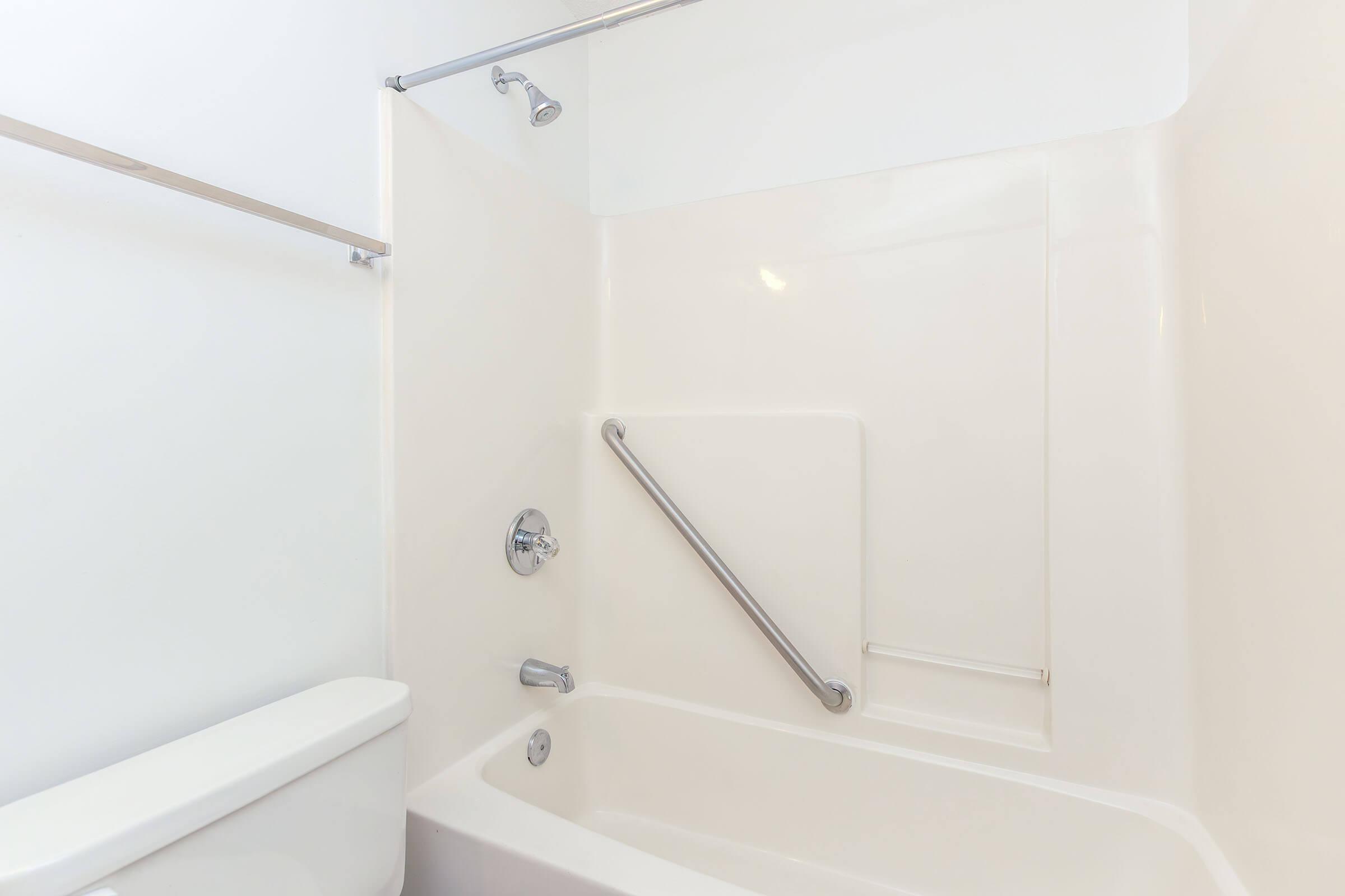 A clean and minimalistic bathroom featuring a white bathtub with a shower head above, grab bar for safety, and a toilet next to it. The walls are plain and bright, contributing to a simple, modern aesthetic.