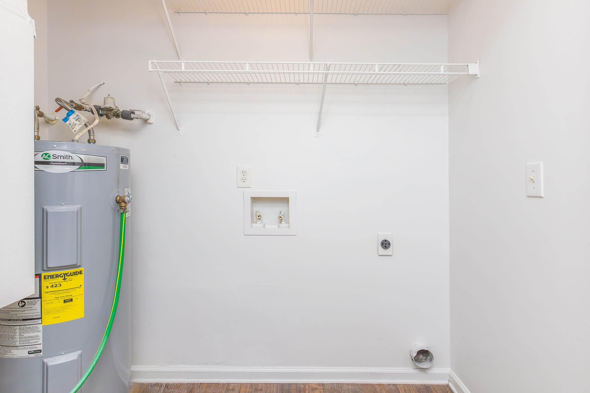 A clean, empty laundry room showcasing a water heater on the left with a green hose attached. There is a white wire shelf above, and the walls are painted white. Electrical outlets are visible on the right side of the wall, along with a small utility connection box. The floor is hardwood.