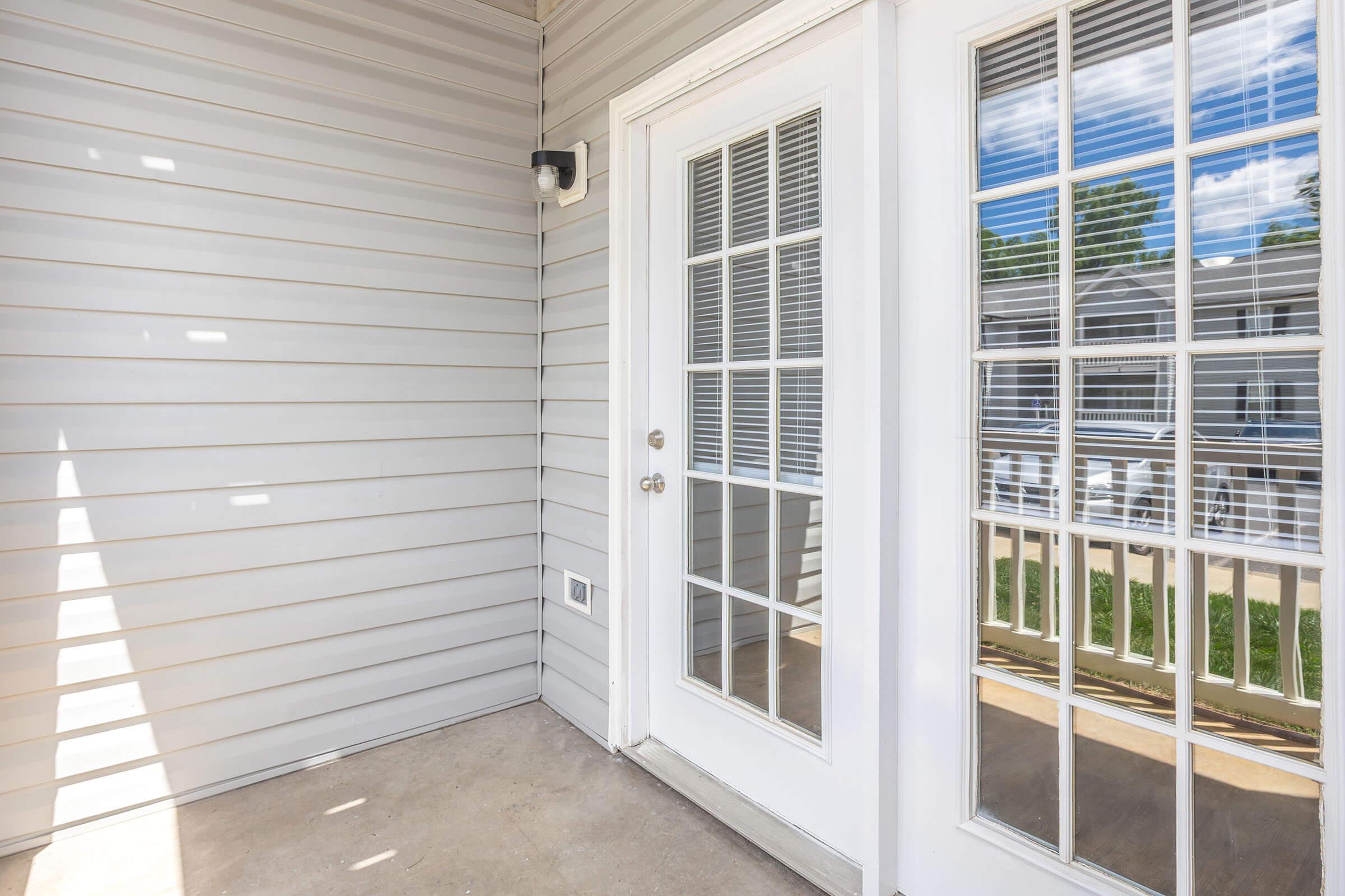 A small patio area featuring a white door with multiple glass panes, leading to a bright, outdoor space. The wall is light grey with horizontal siding, and there is a light fixture above the door. The floor is concrete, and sunlight filters through, creating a warm ambiance.