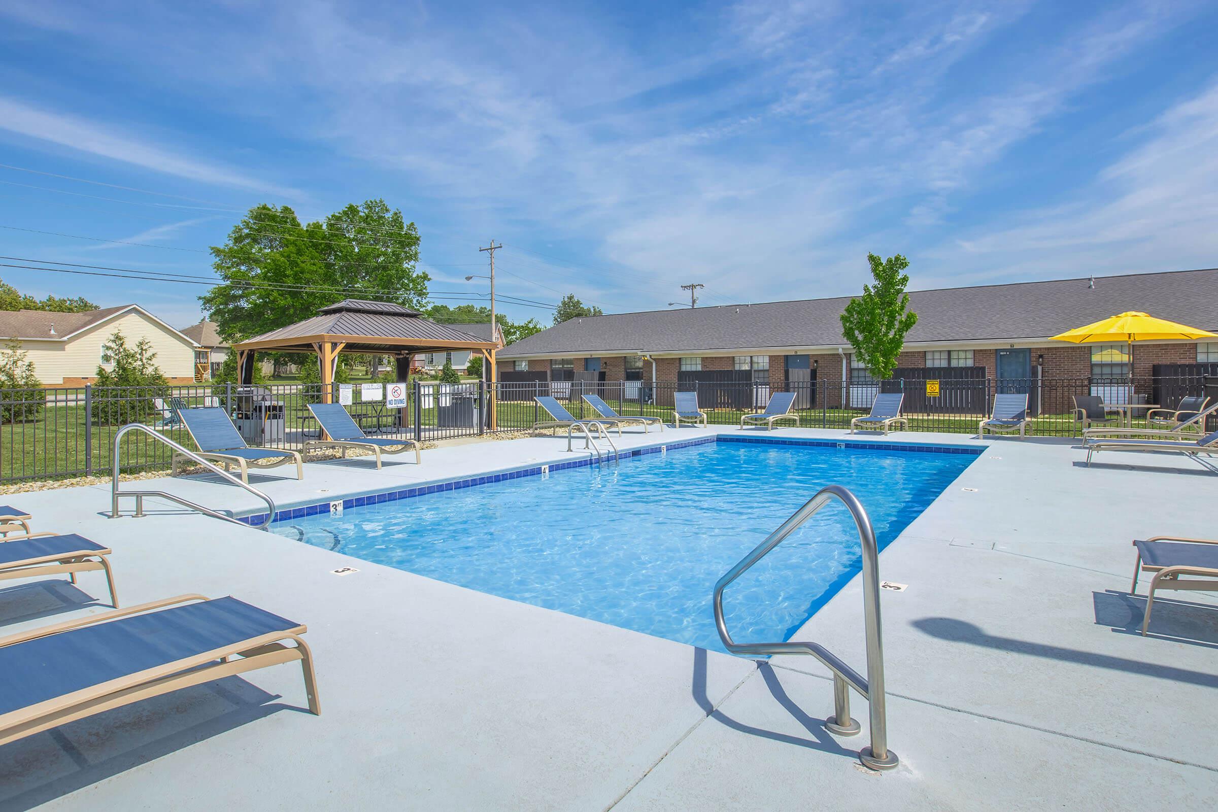 A bright outdoor swimming pool surrounded by lounge chairs and a gazebo. The pool features steps leading into the water, and there are trees and a building visible in the background. The sky is clear with a few clouds, creating a sunny atmosphere for relaxation.