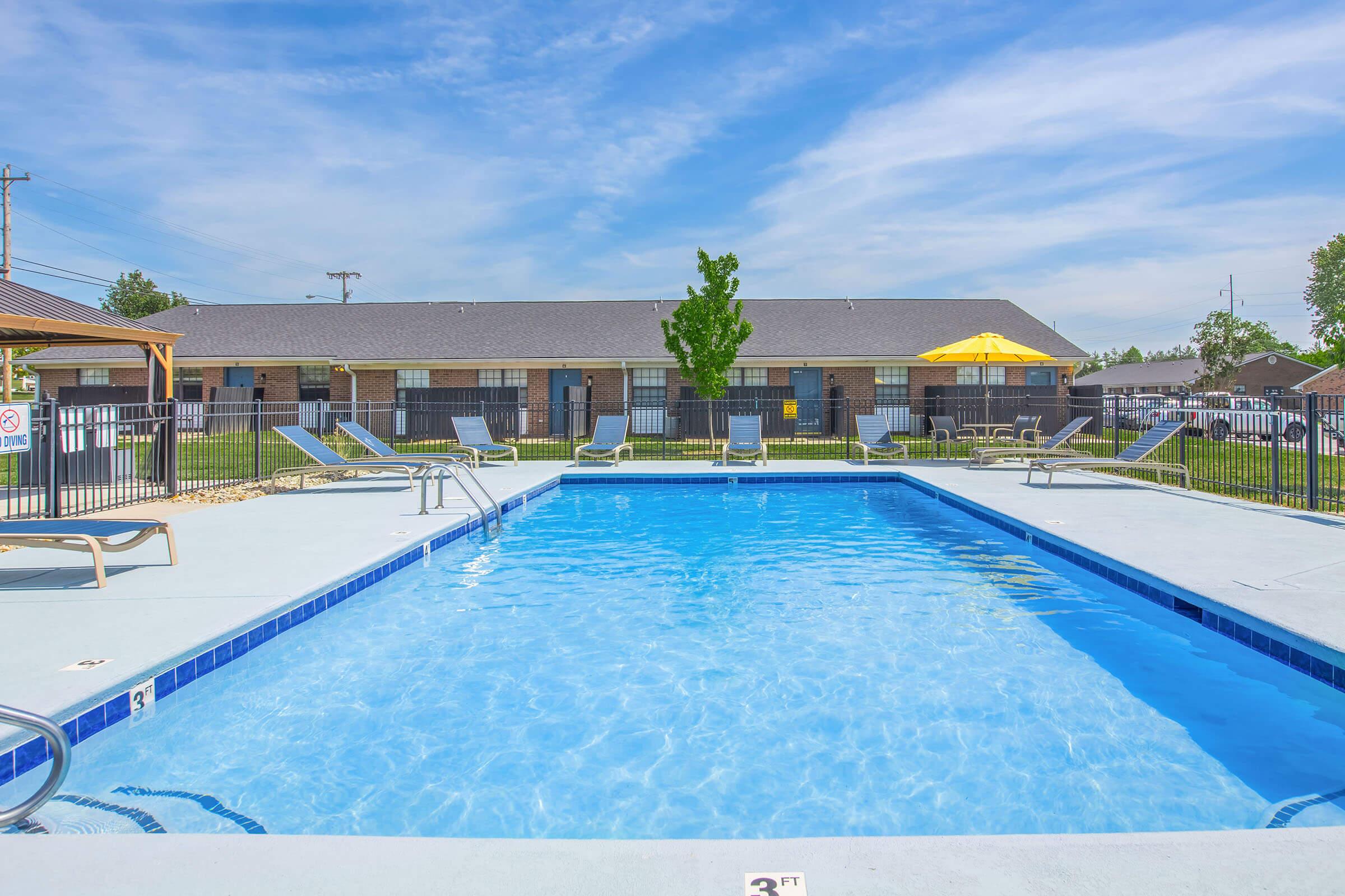 A blue swimming pool surrounded by lounge chairs and a fenced area. There are grassy spaces and buildings in the background, with a bright yellow umbrella providing shade. The sky is clear with some wispy clouds.