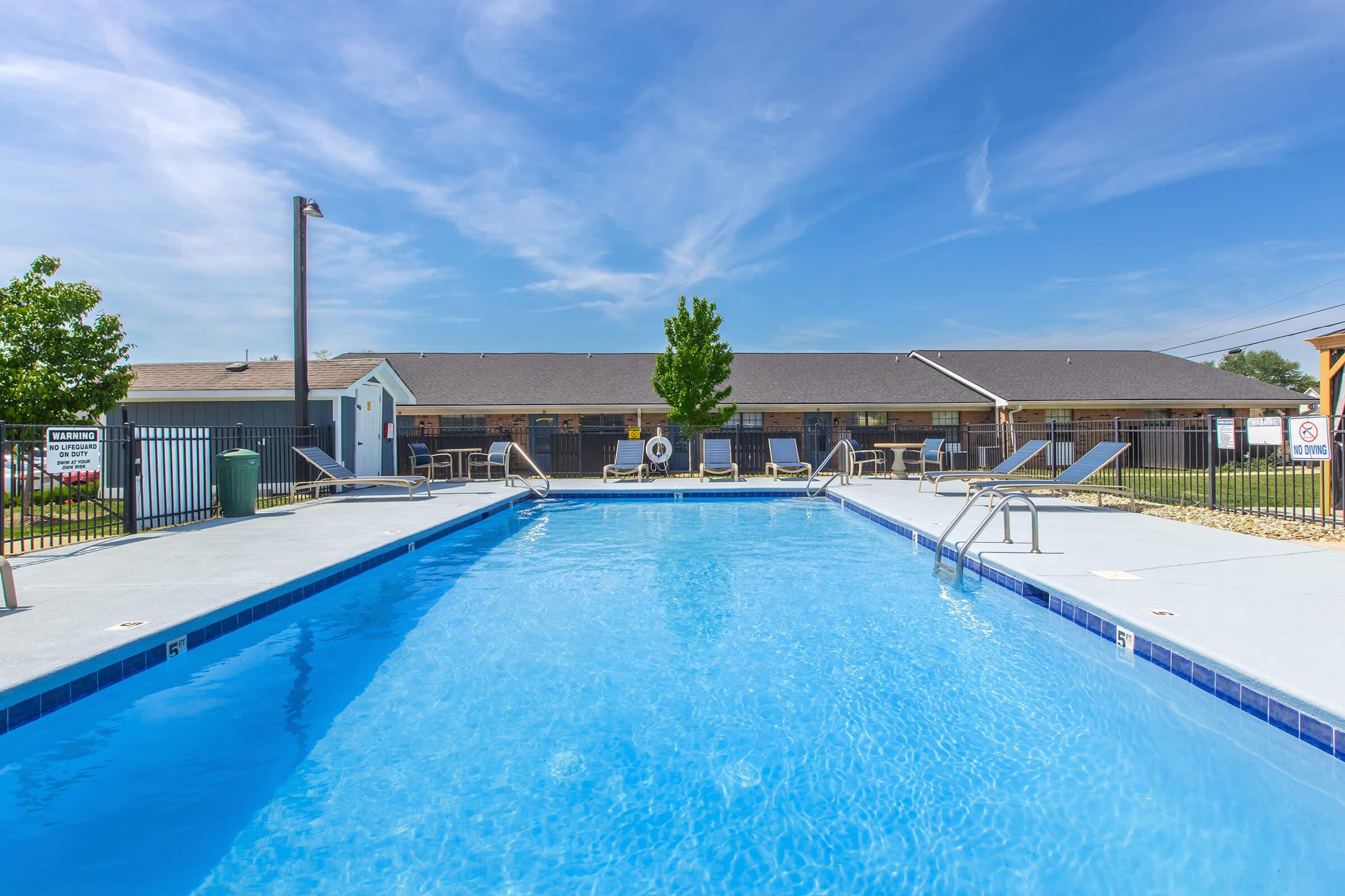 A clear blue swimming pool surrounded by lounge chairs, with a backdrop of a building under a bright sky. The pool area is enclosed with a fence, and there are trees nearby, creating a relaxing outdoor atmosphere.