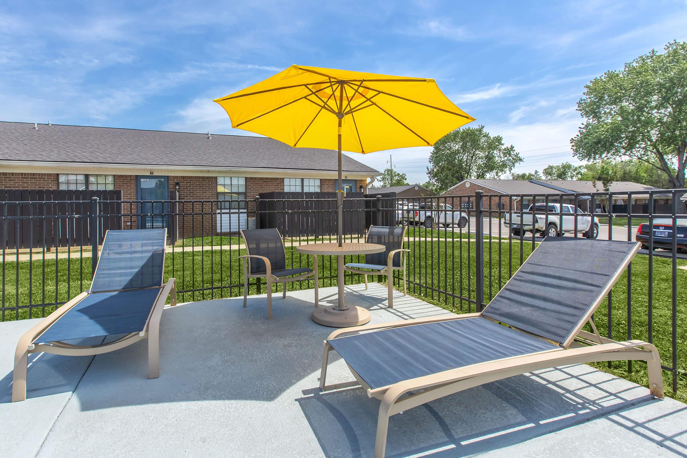 A poolside area featuring two lounge chairs and a round table with a yellow umbrella, surrounded by a black fence. In the background, a residential building and green grass are visible under a clear blue sky.