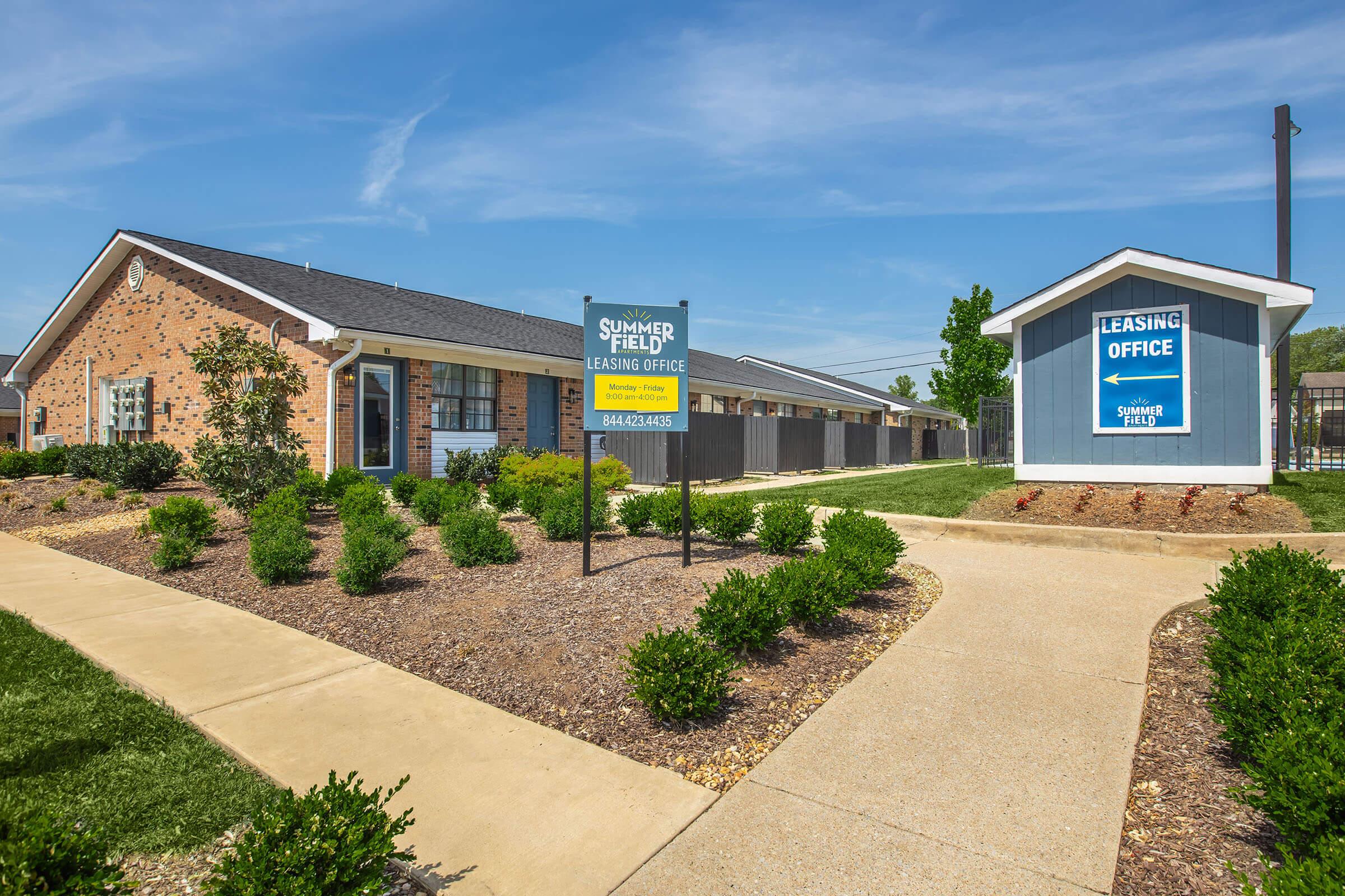 A well-maintained leasing office building at Summerfield, set in a landscaped environment. The structure features a mix of brick and siding, with a prominent sign indicating the leasing office. Neat pathways and greenery enhance the attractive appearance of the property under a clear blue sky.