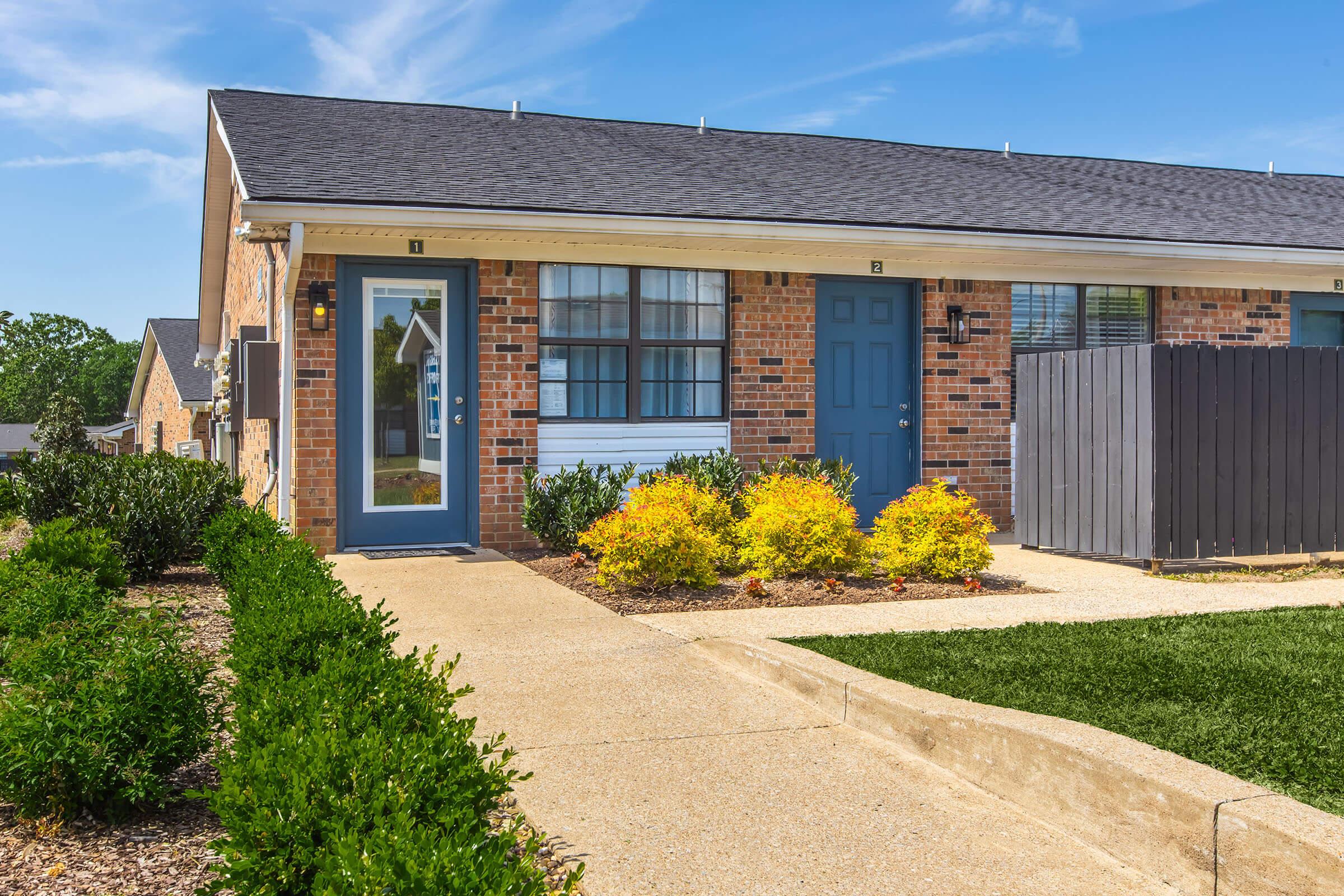 Exterior view of a modern brick townhouse with a blue front door and window. The pathway is bordered by manicured shrubs and colorful plants, leading to the entrance. The sky is clear and blue, creating a bright and inviting atmosphere. A privacy fence is visible beside the house.