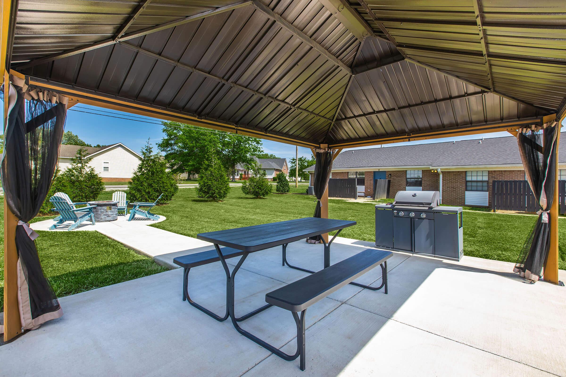 A spacious outdoor patio with a covered gazebo featuring a picnic table and a barbecue grill. Surrounding the area is a well-maintained lawn with neatly arranged Adirondack chairs and small trees, creating a relaxing atmosphere for outdoor gatherings.