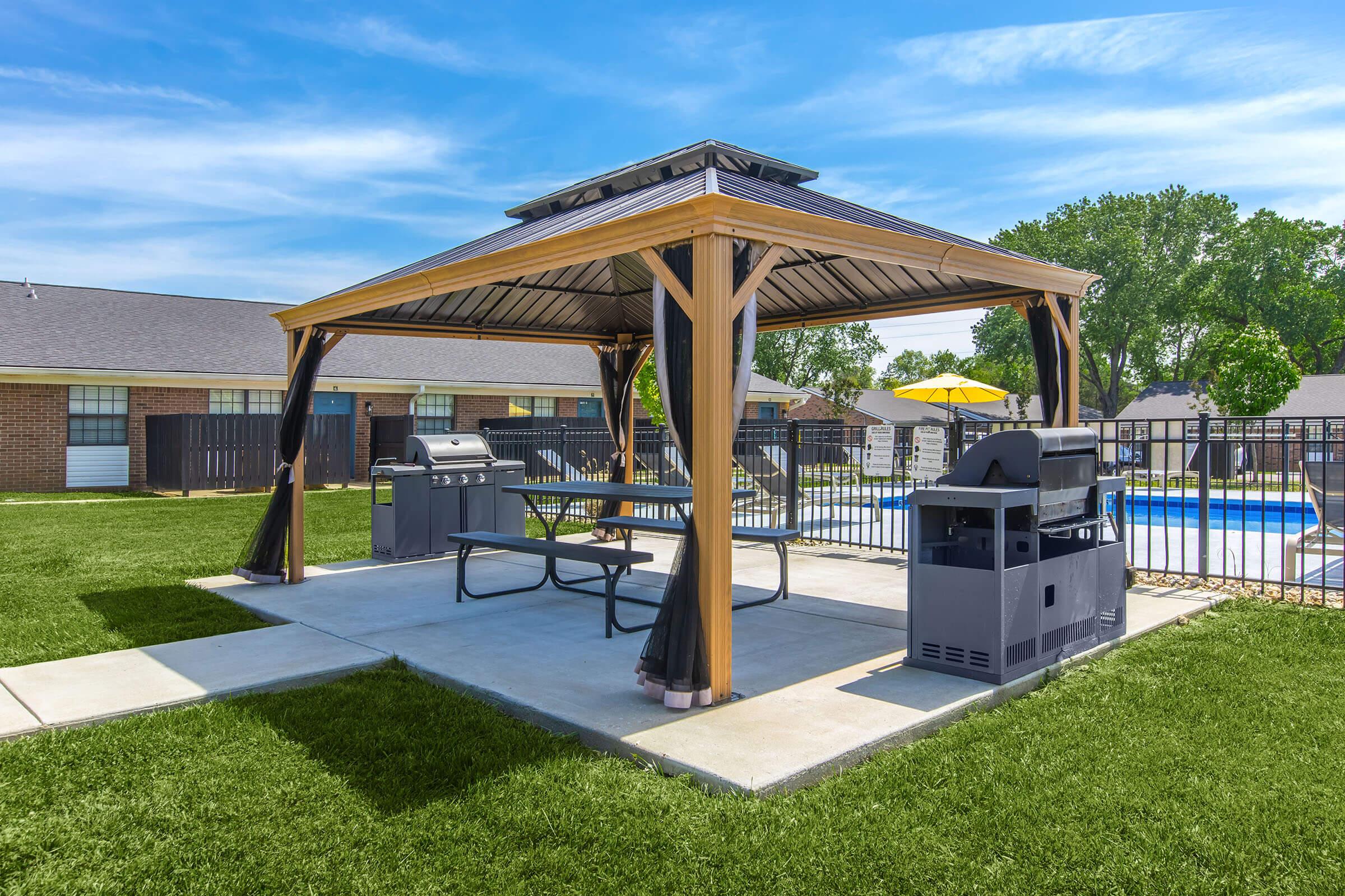 A shaded outdoor gazebo with wooden beams, featuring a picnic table and a charcoal grill. Nearby, another grill is positioned on a concrete slab. The area is surrounded by green grass, and a swimming pool can be seen in the background, along with a sunny blue sky.